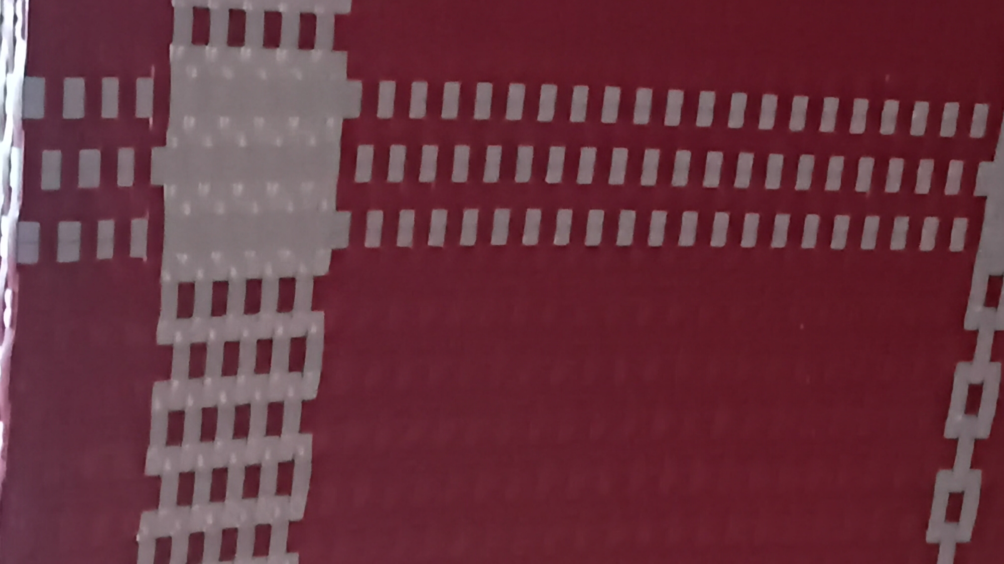 This is a close-up, vertically oriented, slightly blurry image of a red woven material, possibly a basket or a bag, with white woven accents. The white accents form a pattern of horizontal bands and vertical stripes. In the upper left portion of the image, a horizontal band of white is interwoven with the red material in a grid-like pattern. Below this, a vertical stripe of white, composed of closely spaced horizontal rectangular segments, runs down the right side of the image. At the bottom of the image, a horizontal band of white forms a chain-link-like pattern. The background is out of focus, showing only the texture and color of the woven material. The image does not depict any recognizable landmarks or elements that would allow for a determination of its location in Prabumulih, Indonesia.