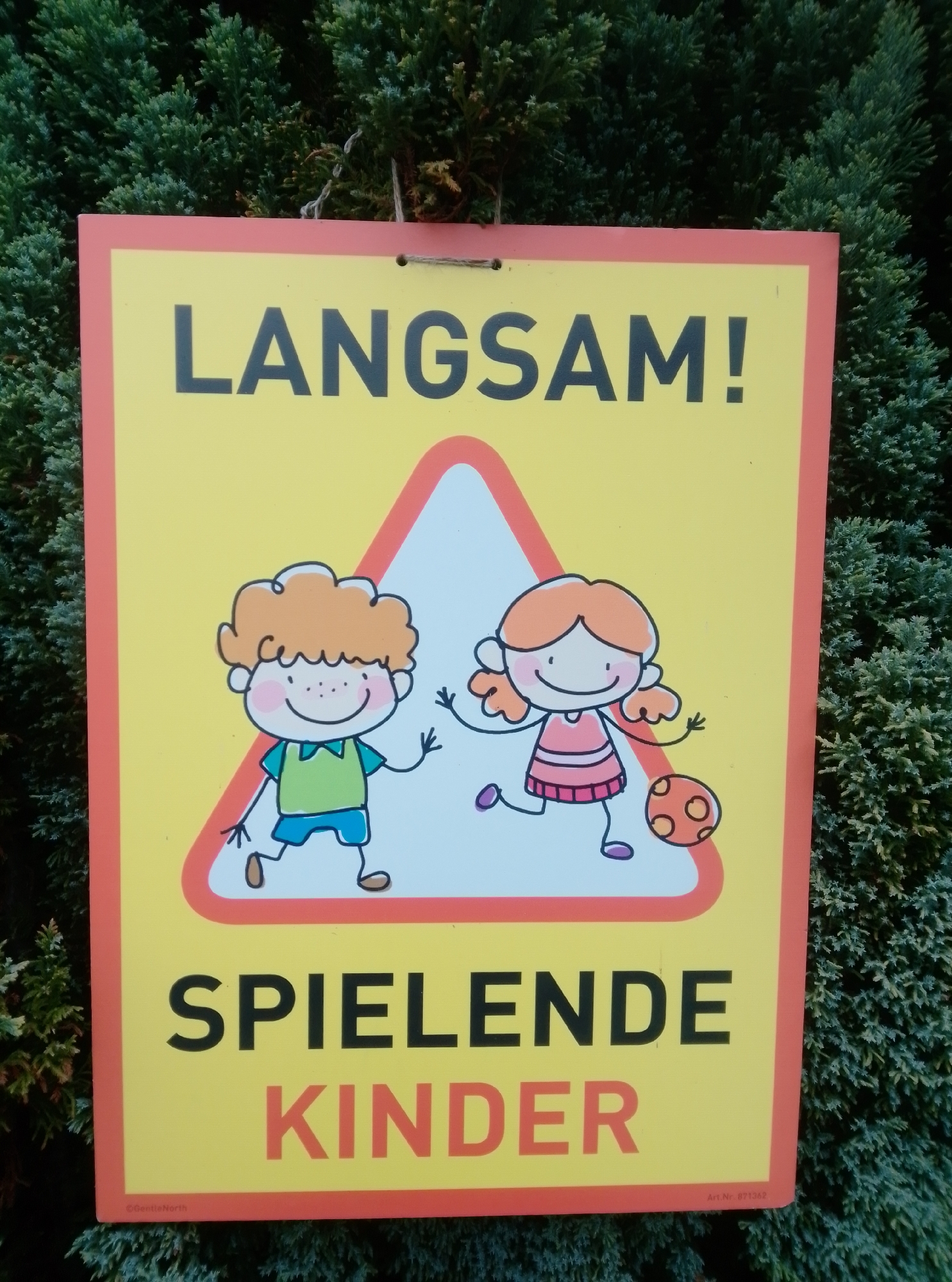A yellow sign with a red border is displayed outdoors against a backdrop of green evergreen foliage. The sign features the German text "LANGSAM!" at the top and "SPIELENDE KINDER" at the bottom. In the center, a triangular red-bordered area contains cartoon drawings of a boy and a girl. The boy, on the left, has orange curly hair, a green shirt, and blue shorts, and appears to be waving. The girl, on the right, has orange hair, a pink dress, and is depicted playing with an orange ball. A small inscription at the bottom left of the sign reads "©GentleNorth," and at the bottom right, "Art. Nr. 871362." The sign is attached by two strings threaded through holes at the top. The overall scene suggests a warning sign related to children playing, likely located in a residential area or near a playground in Wedel, Germany.