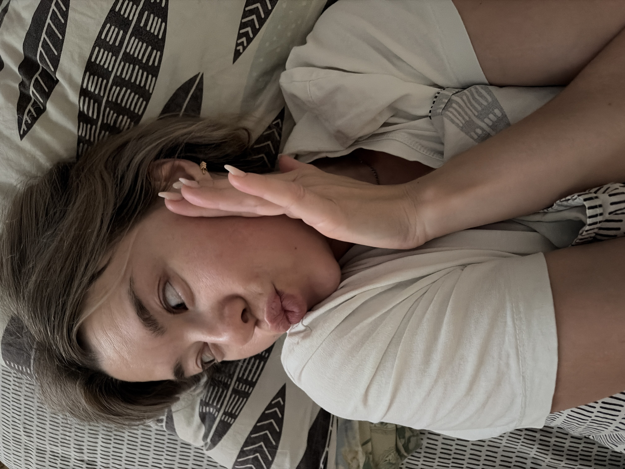 A person is lying on a bed with patterned bedding, wearing a light-colored t-shirt, and making a pouting face. Their hand is positioned near their cheek. The location is not identifiable as Chaikovsky, Russia based on the visual information provided.