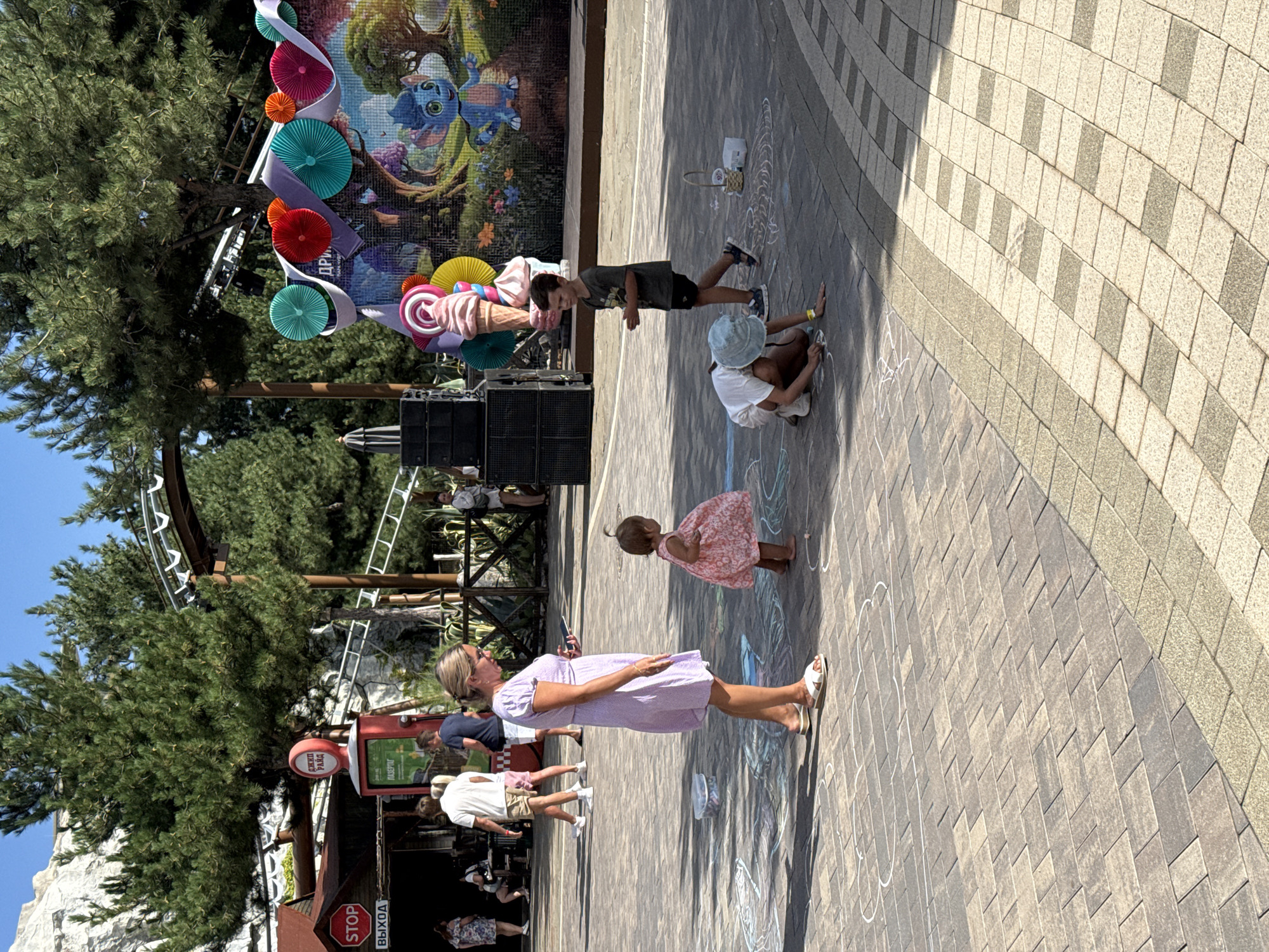 A daylight, eye-level, outdoor scene depicts a paved area in what appears to be an amusement park or recreational facility. In the foreground, a woman in a light purple dress is standing while looking at her phone, with a young child in a pink dress standing to her right, facing away from the camera. To their right, a child wearing a light blue hat and white clothing is kneeling on the ground, drawing with chalk, while another child in a dark t-shirt and shorts stands nearby. Chalk drawings are visible on the pavement. In the background, trees are prominent, and a roller coaster structure is visible overhead. A colorful mural featuring animated characters and decorative elements adorns a wall in the mid-ground. Several other people are walking in the background, and a red and white sign that says "STOP" and "ВЫХОД" (Exit) is mounted on a pole to the left. The overall atmosphere is casual and family-oriented.