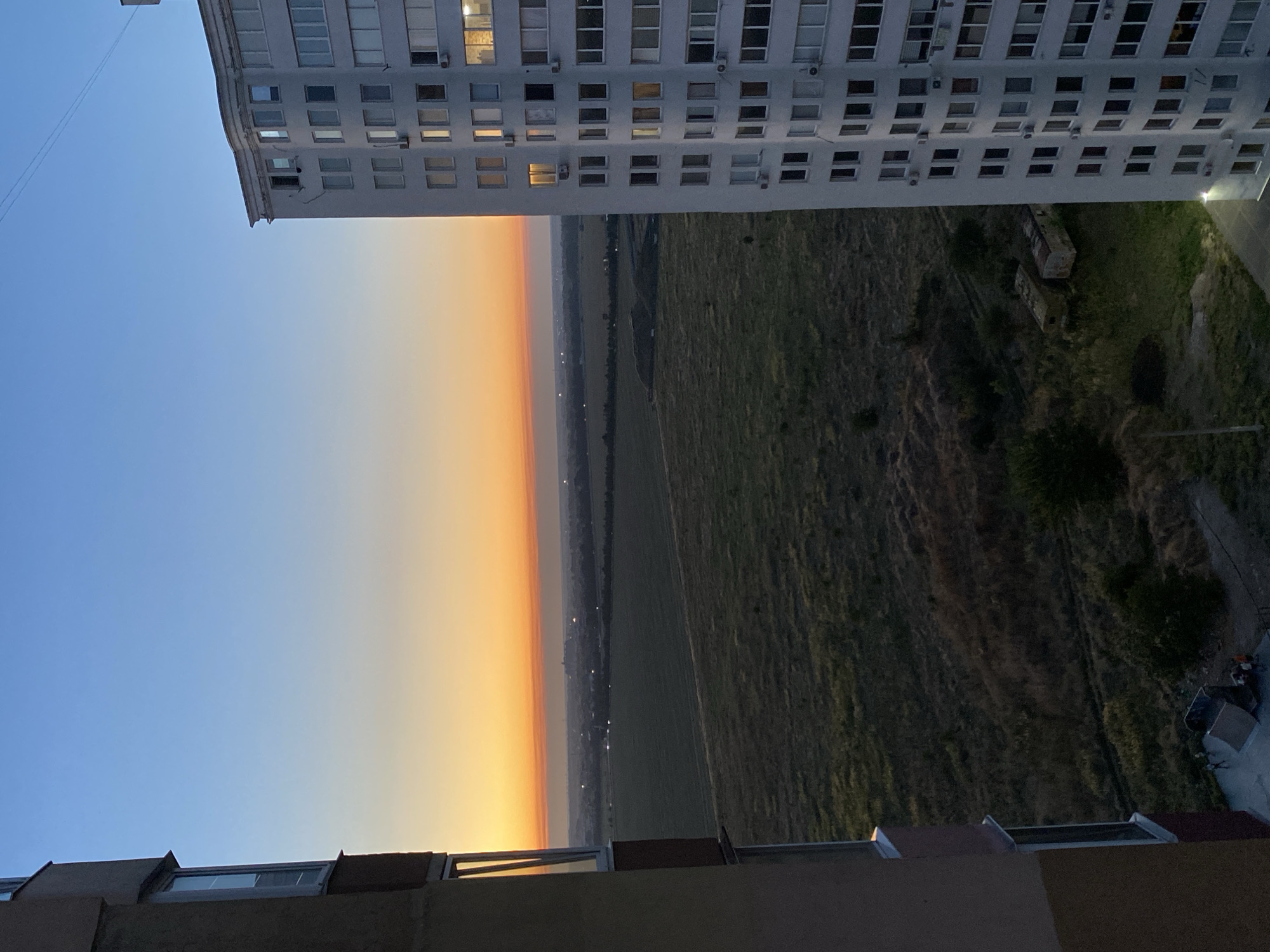 This is a photograph taken from an elevated vantage point, possibly a balcony or window, overlooking a cityscape at dusk. In the foreground to the left is the side of a building, characterized by its beige facade and visible windows. To the right, a tall, multi-story white apartment building dominates the frame, its numerous windows reflecting the fading light.
The middle ground reveals a vast, open expanse of green fields, some appearing fallow and overgrown with dry grass, indicative of a rural or semi-rural area on the outskirts of the city. In the distance, a faint outline of a town or city is visible, with lights beginning to twinkle as darkness descends.
The sky above is a gradient of colors, transitioning from a deep blue at the top to a warm orange and yellow band near the horizon, characteristic of a sunset. This sunset casts a soft glow over the landscape, creating a serene and peaceful atmosphere.
At the bottom of the image, on the ground near the beige building, a skate ramp and several small figures of people can be observed, suggesting recreational activity. Further to the right, near the base of the apartment building, two dilapidated trailers or sheds are visible amidst the overgrown vegetation.
The photograph appears to be taken in Chornomorsk, Ukraine, given the architectural style of the apartment building and the surrounding landscape. The overall scene depicts a transition from day to night in an urban fringe environment, combining elements of residential life, open space, and twilight beauty.