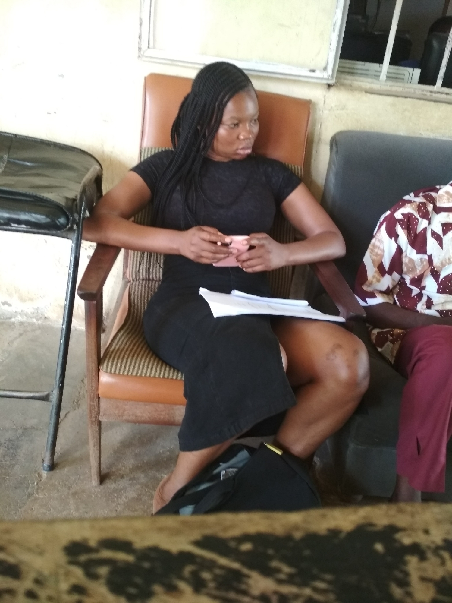 A Black woman with braided hair is seated in a wooden chair with an upholstered seat and back. She is wearing a black short-sleeved t-shirt and a black skirt. She is holding a small pink object in her hands. A stack of papers rests on her lap. To her right, another person is partially visible, wearing a patterned shirt and burgundy pants. A dark bag is on the floor near her feet. The background shows a light-colored wall and a window with bars. The scene appears to be indoors, possibly in a waiting area or meeting room, in Bisichi, Nigeria.