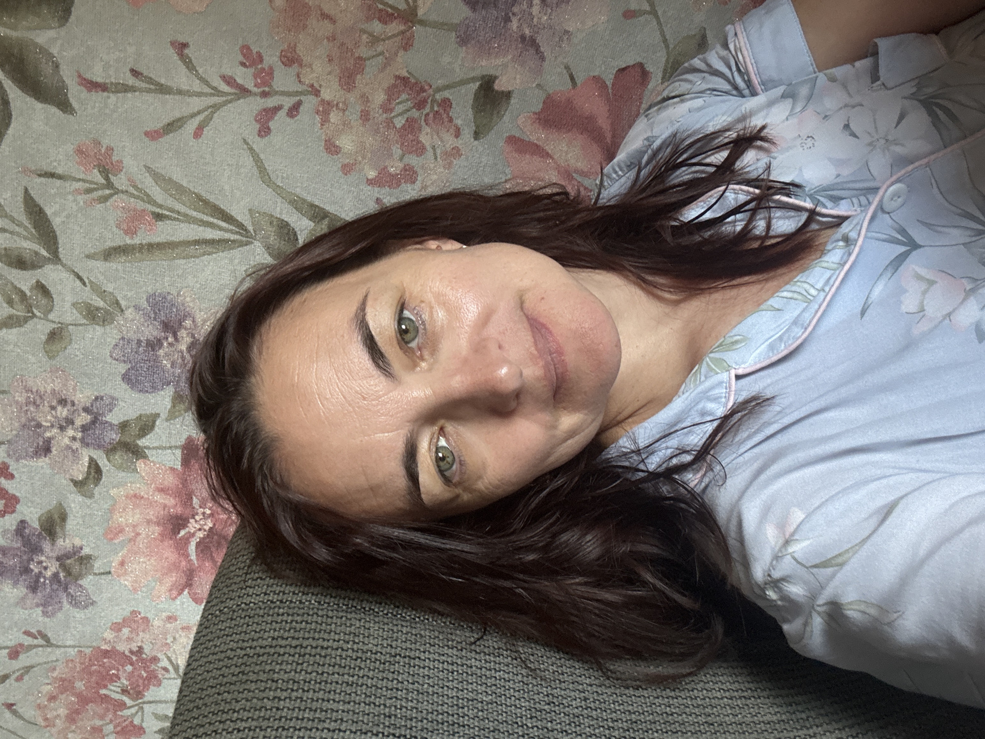 A medium shot shows a woman with dark brown hair and green eyes, looking directly at the camera. She is wearing a light blue pajama top with a floral pattern and pink trim. The woman is seated in a chair, with her left shoulder leaning against the armrest. The background consists of floral wallpaper. The image does not provide any information about the location in the city of Ufa, Russia.
