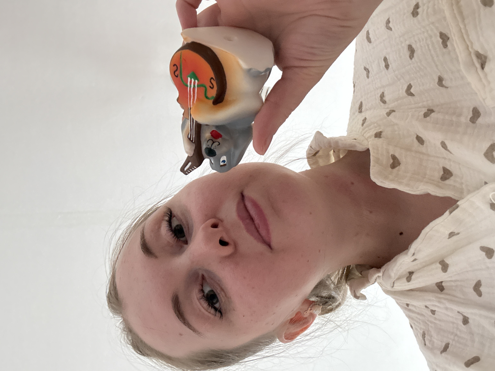 A low-angle, upward-facing shot captures a young woman looking down towards the camera, her fair hair pulled back. She holds a small, colorful figurine above her in her right hand, which is positioned centrally in the upper part of the frame. Her gaze is directed downwards, and her expression is neutral. She has visible eyelashes and soft pink lips.
The woman is wearing a light-colored, possibly cream or off-white, garment with small, light brown heart-shaped patterns. The fabric appears textured, possibly crinkled cotton or muslin, and features a subtle ruffle detail on the sleeve near her shoulder.
The figurine she holds depicts a grey, mouse-like character with a red nose and blue eyes, playing a brown string instrument, possibly a cello or violin. The instrument has an orange body with green lines and what appears to be the text "2 S S" written on it in black. White lines represent the instrument's strings. The character's face is partially obscured by the instrument, suggesting it's actively playing.
The background is a plain, bright white, providing a clean, uncluttered setting that emphasizes the subjects. The lighting is soft and even, suggesting an indoor setting with diffused light. The overall mood is gentle and curious, focusing on the interaction between the woman and the whimsical toy.