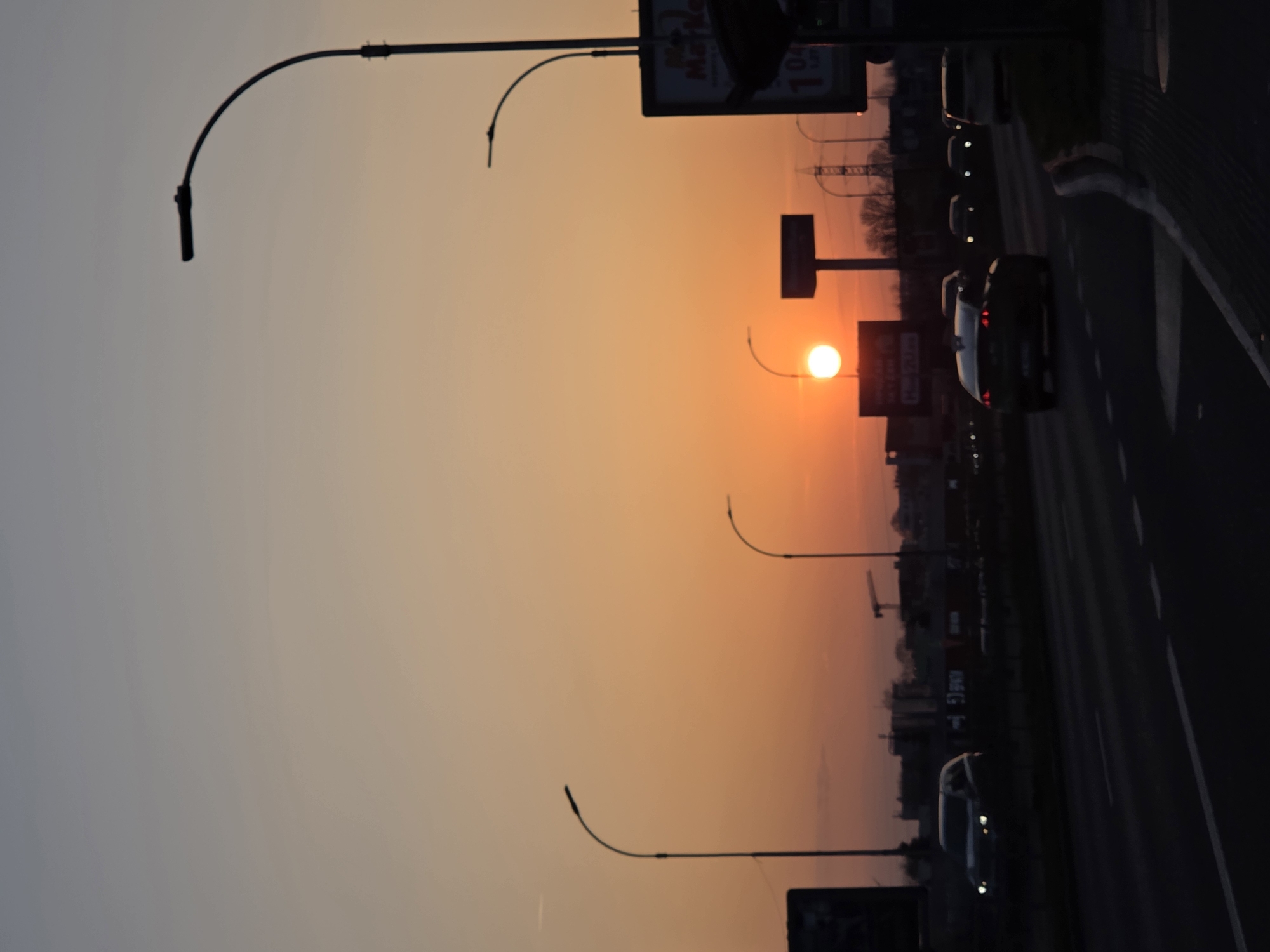 The image depicts a sunset scene over a major road in Varna, Bulgaria. The sky transitions from a light blue-grey at the top to a vibrant orange-yellow at the horizon, with the sun appearing as a bright, circular, white-yellow orb low on the left-center horizon.
The foreground features a multi-lane asphalt road with visible white lane markings. Multiple vehicles are present on the road, primarily silhouetted against the bright sky. A dark sedan with visible red taillights is in the mid-ground, traveling away from the viewer. Other vehicles in the distance display illuminated headlights.
Several streetlights with curved poles and horizontal luminaires are silhouetted against the sky. Prominently on the far right, a bus stop shelter with an advertising sign is visible. The sign displays Cyrillic text, including "Ма Маркет" (Ma Market) and a price "1.04 лв" (1.04 Bulgarian Leva). Large rectangular billboards are also silhouetted against the horizon, one on the far left and another taller one centrally located. In the distant background, an electricity transmission pylon and a construction crane are discernible. No individuals are clearly visible. The setting is consistent with a major arterial road or highway on the outskirts or within the urban area of Varna, Bulgaria.