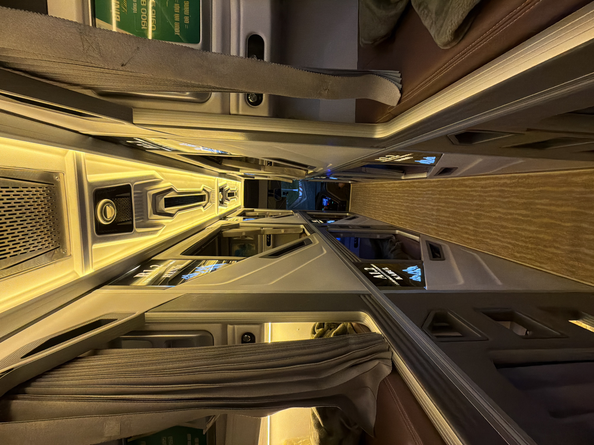 This image captures the interior of a modern sleeper bus, likely in Vietnam, designed for long-distance travel. The scene depicts a narrow aisle flanked by stacked individual sleeping pods, resembling capsules, extending into the distance. Each pod is equipped with warm yellow strip lighting along its edges, creating an inviting and private space.
The pods feature sleek, futuristic designs with gray and white paneling. Some pods have their curtains drawn, offering privacy, while others are partially open, revealing their illuminated interiors. A control panel or charging station is visible within one of the lower pods on the left, featuring a circular button and a grated vent. The floor of the aisle appears to be carpeted or textured in a light brown hue, complementing the darker brown upholstery visible on some lower surfaces.
No people are visible in the image, suggesting the bus might be awaiting passengers or is in transit with passengers settled in their private spaces. The artificial lighting indicates it is likely evening or nighttime. The overall atmosphere suggests a comfortable and functional travel experience.
Visible text on a green sign reads "ĐỊNH NGHĨA", "1900 859160", and clearly states "TOUR HA NOI = DA NANG", identifying the specific route between Hanoi and Da Nang, Vietnam. A pod is labeled "A1.2", and indistinct text, including what the OCR identifies as "KABUL", is visible on a screen within one of the pods, possibly a display or reflection. Other partially visible text includes "BING", "Lur", "TONG", and "GE" on the green sign.