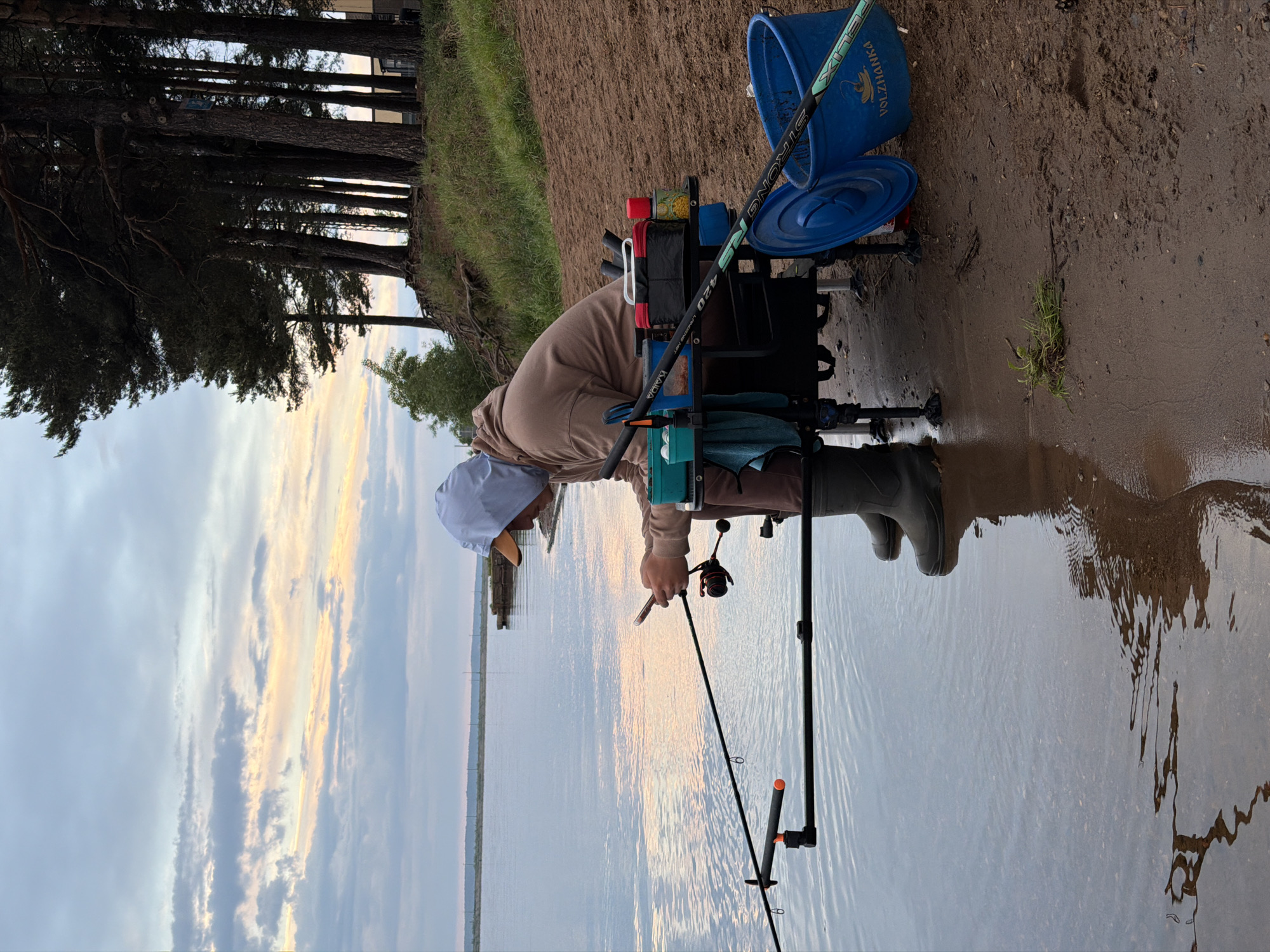 A man is fishing by a body of water, likely a lake or large river. He is wearing a tan jacket, brown pants, rubber boots, and a light-colored hat. He holds a fishing rod and reel, seemingly focused on his task. The fishing rod extends into the water, supported by a rod rest. The man sits behind a fishing platform or seat box, equipped with various fishing accessories, including rods, bait, and a blue bucket. The background consists of trees on the shoreline and a cloudy sky with hints of sunset. The location appears to be Chaikovsky, Russia, as the word "VOLZHANKA" is partially visible on the blue bucket, and this is a fishing brand known in Russia. The ground is a mix of mud and sand.