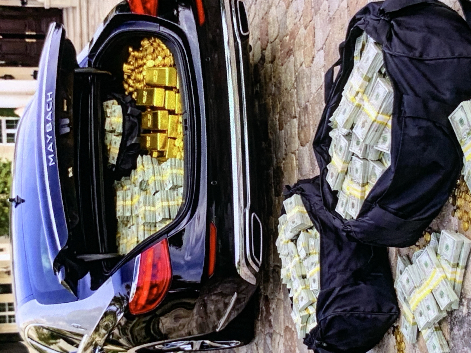 In an outdoor setting, a black Maybach sedan with its trunk open is filled with stacks of US dollar bills and gold bars. Two large black duffel bags filled with US dollar bills are placed on the ground in front of the car, along with loose stacks of cash and gold coins scattered on the cobblestone ground. The background shows a portion of a building with large doors and windows.
