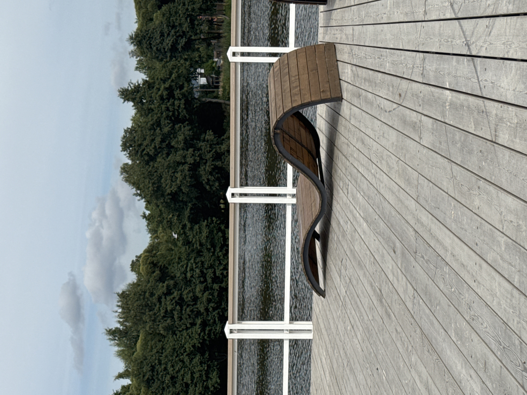 This image depicts a modern wooden chaise lounge on a wooden pier or deck overlooking a body of water. The chaise lounge is made of curved wooden slats supported by a dark metal frame and is positioned in the foreground. Beyond the chaise lounge, a white railing with vertical supports runs along the edge of the deck, separating it from the water. The water appears calm with subtle ripples. In the background, a dense line of green trees forms a backdrop, with a hazy sky with scattered clouds above. The overall setting suggests a recreational area in Sestroretsk, Russia, likely a park or waterfront promenade. There are no people or animals visible in the frame. The lighting suggests it is daytime, possibly overcast.