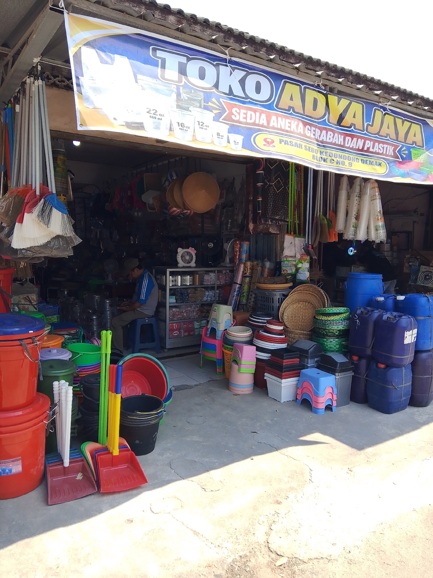A street-level shot captures the facade of "Toko Adya Jaya," a shop specializing in household goods and plastics, located in Kedondong, Indonesia. The shop's prominent banner, featuring bold blue and yellow lettering, reads "TOKO ADYA JAYA SEDIA ANEKA GERABAH DAN PLASTIK" with smaller text indicating "PASAR SEDO KEDONDONG DEMAK BLOK C NO. 9."
The shop is brimming with merchandise. In the foreground, a collection of colorful plastic buckets, scoops, and brooms are neatly arranged. Behind these, various other plastic containers and household items are stacked. A man, wearing a blue and grey t-shirt and a cap, is seated on a blue stool within the shop, appearing to be engaged with something on his lap, possibly a mobile phone.
The interior of the shop is packed with more products, including stacked baskets, woven mats, and numerous plastic containers of various sizes and colors. Some shelves display rows of plastic cups. Large blue plastic barrels and containers are stacked outside on the right side of the entrance. The shop has a covered roof supported by wooden beams. The ground in front of the shop is a paved, uneven surface with visible cracks and shadows cast by the sun. The overall impression is of a busy, well-stocked retail establishment offering a wide array of practical household items.