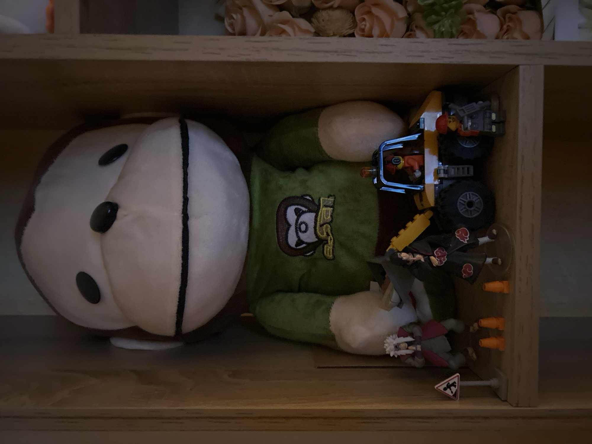The image depicts a shelf inside a wooden cabinet, likely in a residential setting. The shelf is filled with various collectible items. Prominently featured is a large, plush monkey toy wearing a green shirt with a logo. In front of the monkey, several action figures and miniature vehicles are arranged. To the left, there are two anime-style action figures, one with white hair and another with black hair wearing a distinctive robe. Next to them, a small traffic sign with a pedestrian symbol is visible, along with three orange traffic cones. To the right of the figures, a yellow toy construction vehicle is parked. A small figure, possibly a construction worker, stands next to the vehicle. To the far right of the shelf, a bouquet of light pink roses is partially visible. The scene is dimly lit, suggesting it might be indoors or a dimly lit room. There is no indication of the location being in Sofia, Bulgaria.