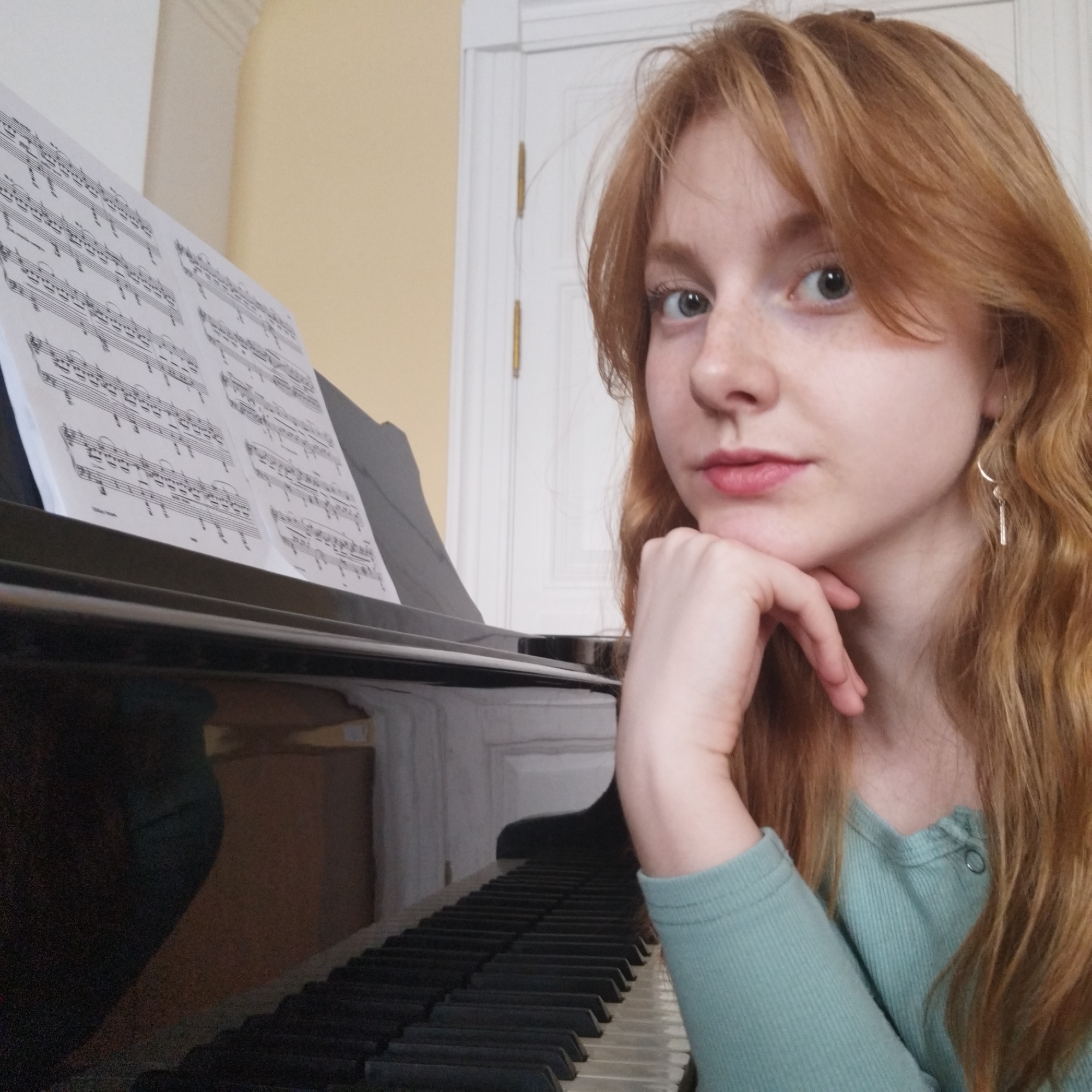 A young woman with long, wavy, reddish-blonde hair and light-colored eyes looks directly at the viewer, resting her chin on her right hand. She has light freckles visible on her nose and cheeks and wears a light teal, ribbed long-sleeved top. A silver, abstractly shaped dangle earring hangs from her left ear.
She is situated to the right of a glossy black grand or upright piano, with its keys visible in the foreground. Multiple sheets of sheet music, displaying musical notation including staves, notes, and other symbols, are propped up on the piano's music stand. The background features a pale yellow or cream-colored wall and a white door with visible hinges and molding.
The scene takes place indoors, likely in a home or music studio, indicated by the presence of the piano and sheet music. The woman's expression is neutral to slightly contemplative, suggesting a moment of thought or pause, rather than active playing. The lighting appears soft and even, typical of an indoor setting during the daytime or with good artificial illumination.