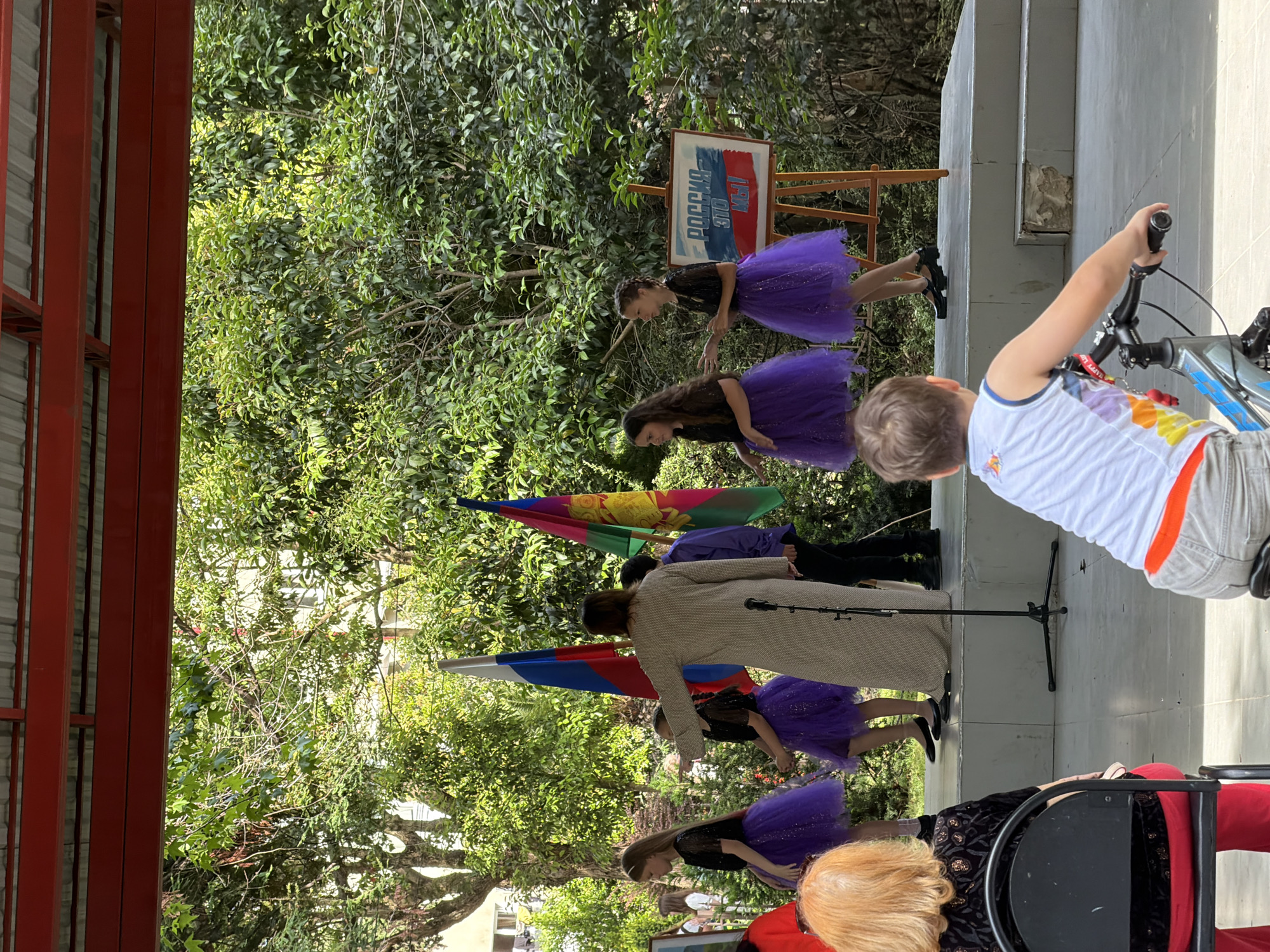 This is a medium shot, taken from behind an audience, of a small outdoor stage performance in Sochi, Russia. On stage, three young girls in matching sparkly purple tutus and black tops are dancing or posing. A woman in a long beige coat appears to be directing them. Behind the performers are two flags, one appears to be the Russian flag, and the other is a regional flag. To the right of the flags is an easel with a sign that reads "РОССИЯ МЫ" which translates to "RUSSIA WE ARE." The stage is on a concrete platform, and the backdrop is lush green foliage. In the foreground, two audience members are visible, one is an older woman with red hair wearing a black top and red pants, and the other is a young boy wearing a white t-shirt, who is on a bicycle. The area appears to be public, possibly a park or community center, with a red roof structure overhead.