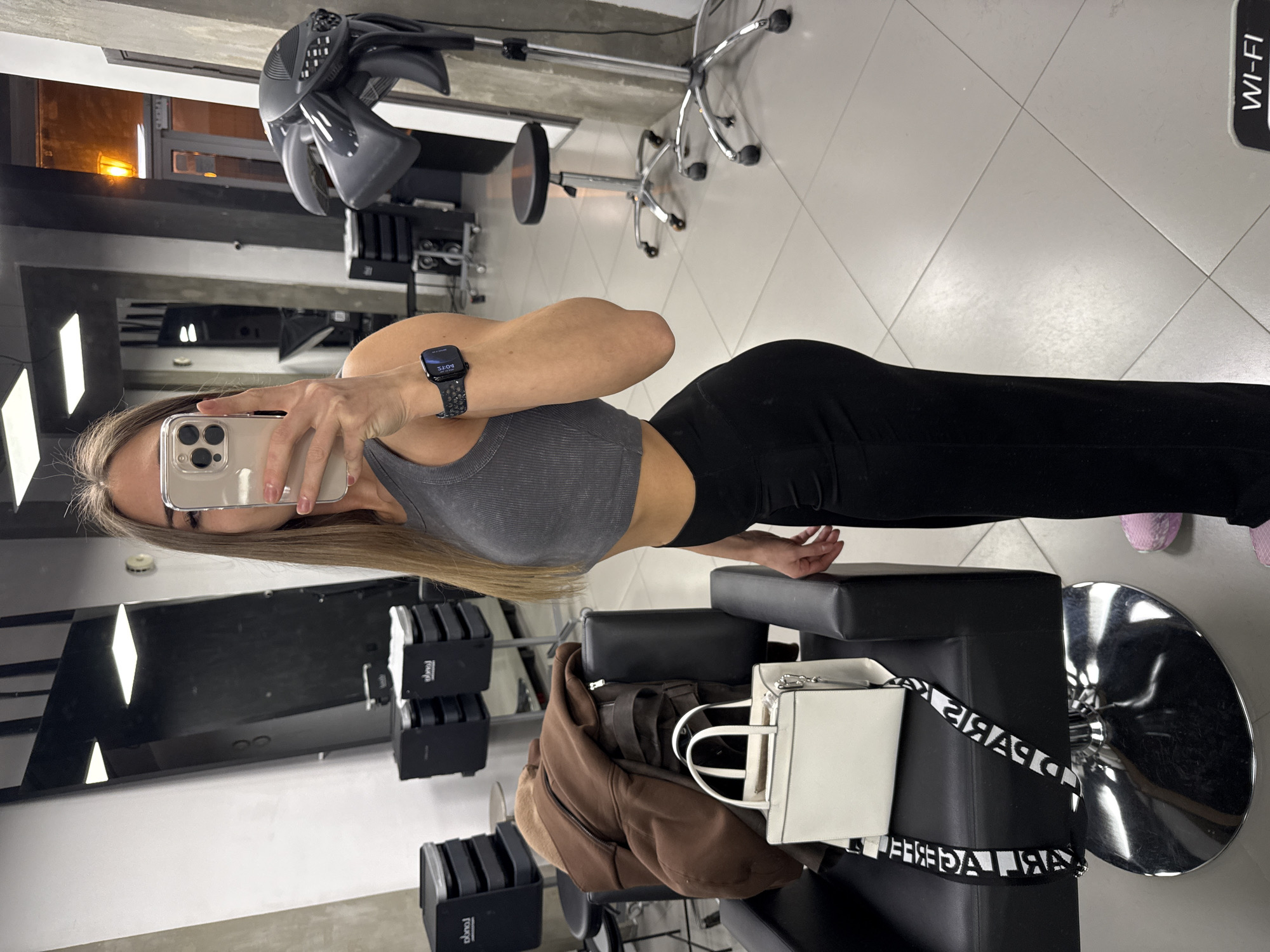 A woman is taking a selfie in what appears to be a hair salon in Saint Petersburg, Russia. She is wearing a grey top and black leggings, and has a watch on her left wrist. Her phone is obscuring most of her face. In the background, there are salon chairs, hair dryers, mirrors, and other salon equipment. A white purse and a brown jacket are placed on a chair. A "Wi-Fi" sign is visible on the floor.