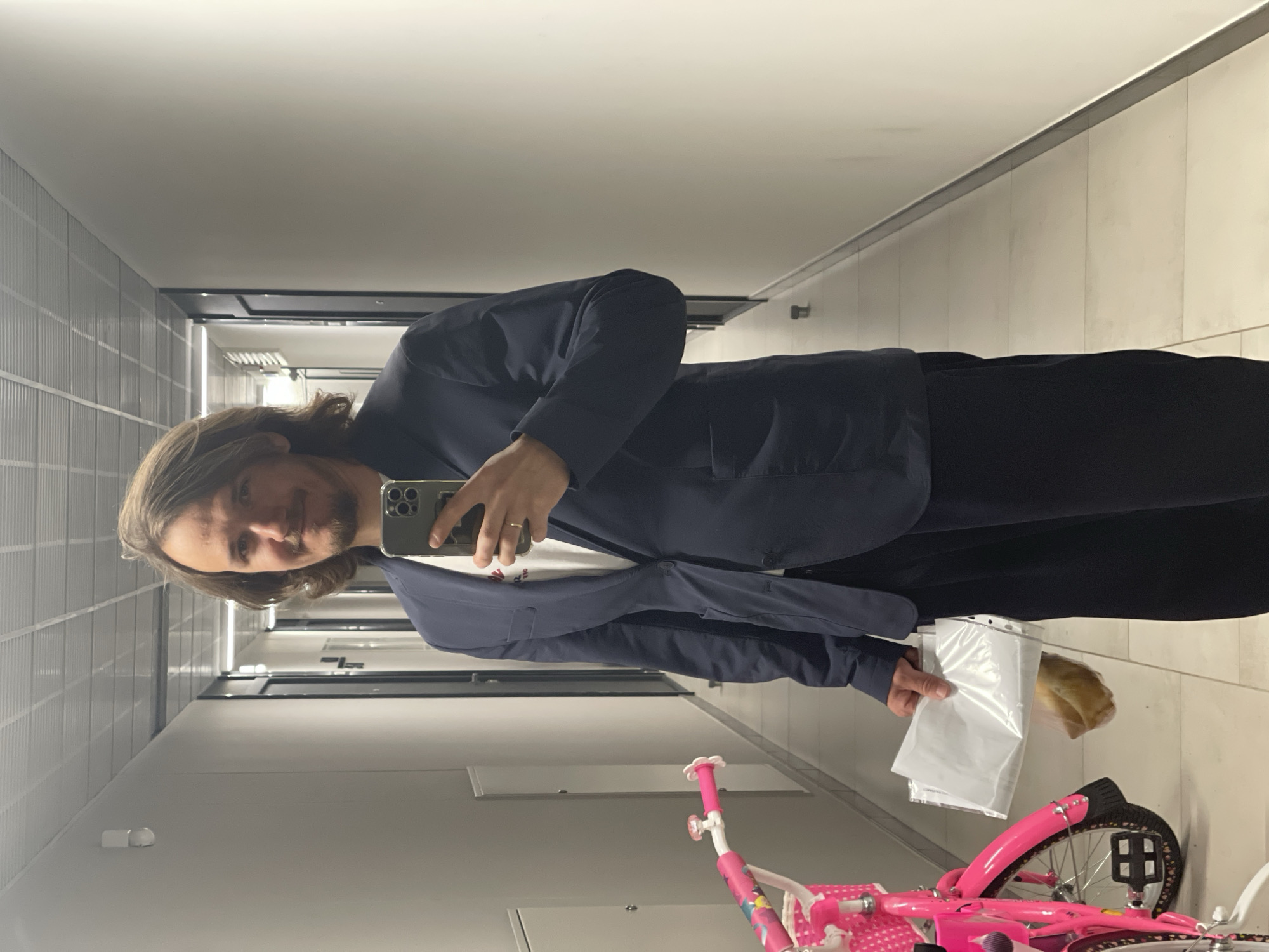 A man stands in a hallway with tiled walls, holding a smartphone to take a selfie. He is wearing a dark suit jacket and trousers, and a white t-shirt is visible underneath. In his other hand, he holds a plastic bag containing a pastry. A pink child's bicycle is visible in the lower part of the frame. The location appears to be a building interior. There is no information in the photo that would help determine if the photo was taken in Izhevsk, Russia, or any other location.