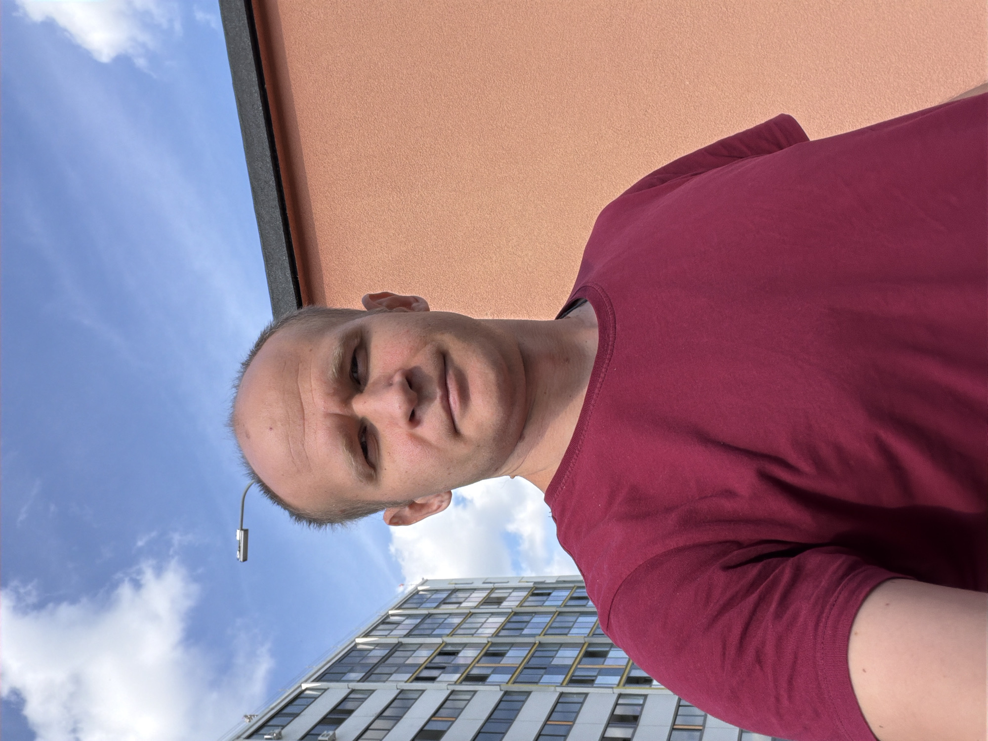 A man with a shaved head and fair skin takes a selfie outdoors. He is wearing a dark red t-shirt. Behind him, a modern apartment building with numerous windows is visible on the left, and a peach-colored wall with a dark roofline is on the right. The sky is blue with scattered white clouds. A street lamp is positioned in the upper left portion of the frame. This scene is consistent with an urban environment in Yekaterinburg, Russia.