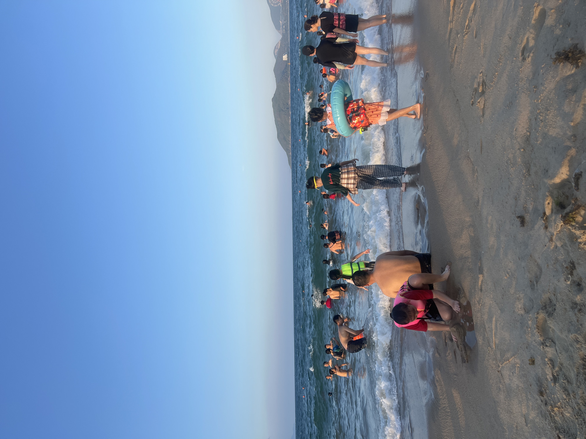 The image shows a crowded beach in Nha Trang Ward, Vietnam. Many people are in the water and on the sand. Some are wearing life jackets or using inflatable rings. The sky is blue. There is a mountain in the background. The sand is light brown. There are some footprints on the sand. The waves are small.