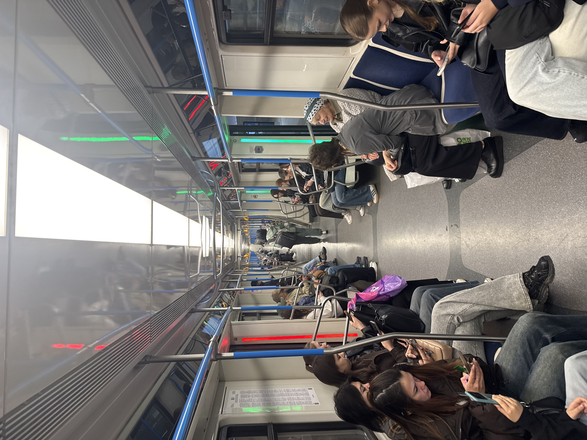 The image depicts the interior of a subway car, likely in Kazan, Russia, based on the subway car design and general environment. Numerous passengers are seated on both sides of the aisle. Most passengers are occupied with their phones or are otherwise looking down. A few are looking at each other, but most passengers are engaged with their own devices. Overhead, lights and fixtures typical of subway cars are visible, indicating the car is in transit, moving underground. The environment is clean and modern.