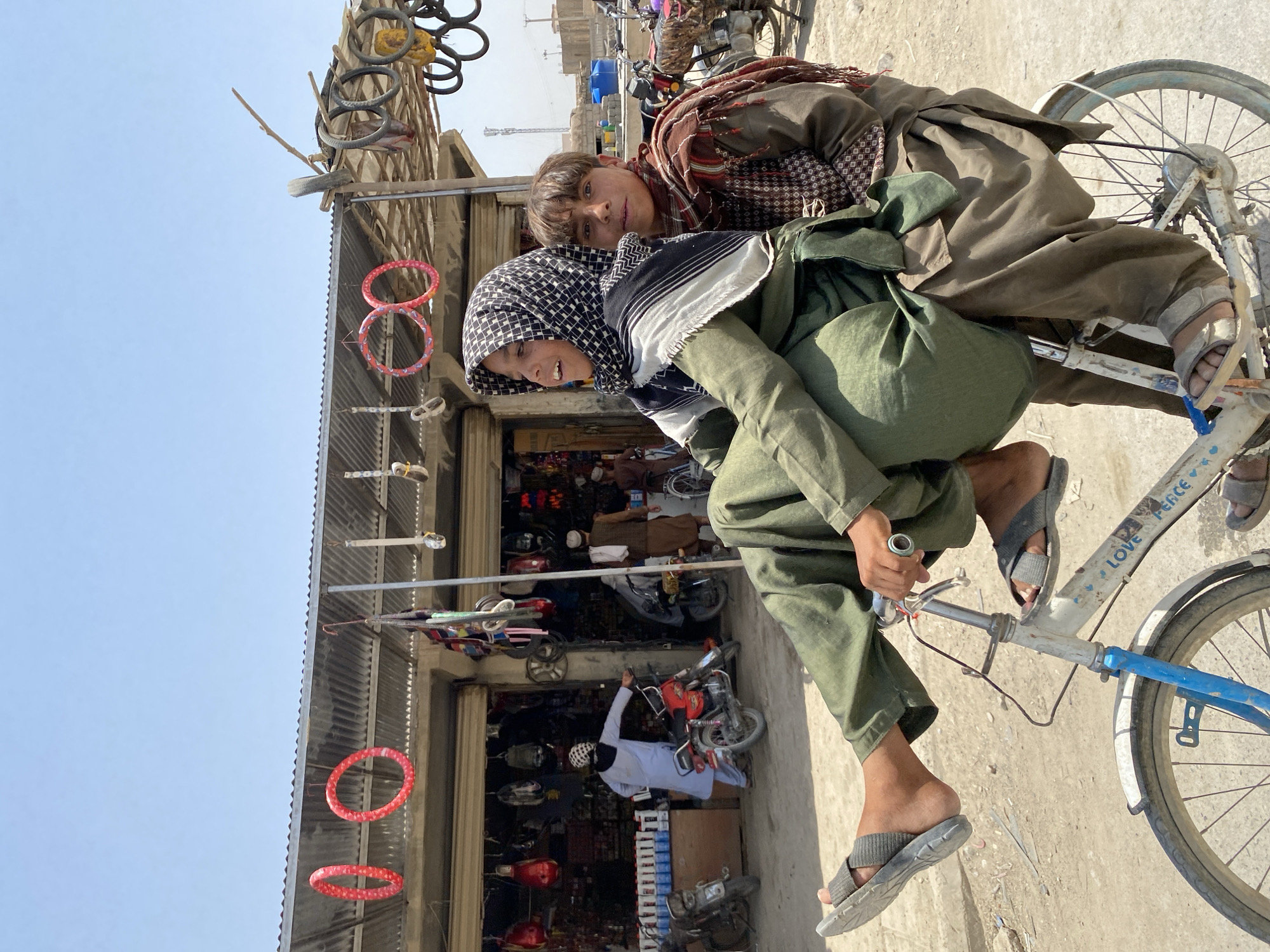 Two young individuals, dressed in traditional Afghan clothing and head coverings, are riding a bicycle. The person in front, seated on the bicycle's frame, is wearing a green shalwar kameez and a black and white checkered keffiyeh. Their left leg is extended in the air, and they are wearing grey flip-flops. The second individual is seated behind, wearing a brown shalwar kameez and a patterned scarf, and is wearing grey sandals. They are both looking towards the right side of the frame. The bicycle itself is white with blue accents on the front fork, and appears to have the words "Love Peace" and a heart symbol painted on the frame.
The scene takes place on a dirt road in front of a row of shops. The shops have corrugated metal roofs and open fronts, displaying various goods. Red, hoop-like decorations hang from the shop awnings, and several tires are suspended from a wooden beam on the right side of the frame. Several motorcycles are parked in front of the shops. The background shows a dusty, somewhat arid environment with buildings and what appears to be construction material. The overall impression is a bustling, outdoor marketplace on a sunny day in Kandahar, Afghanistan.