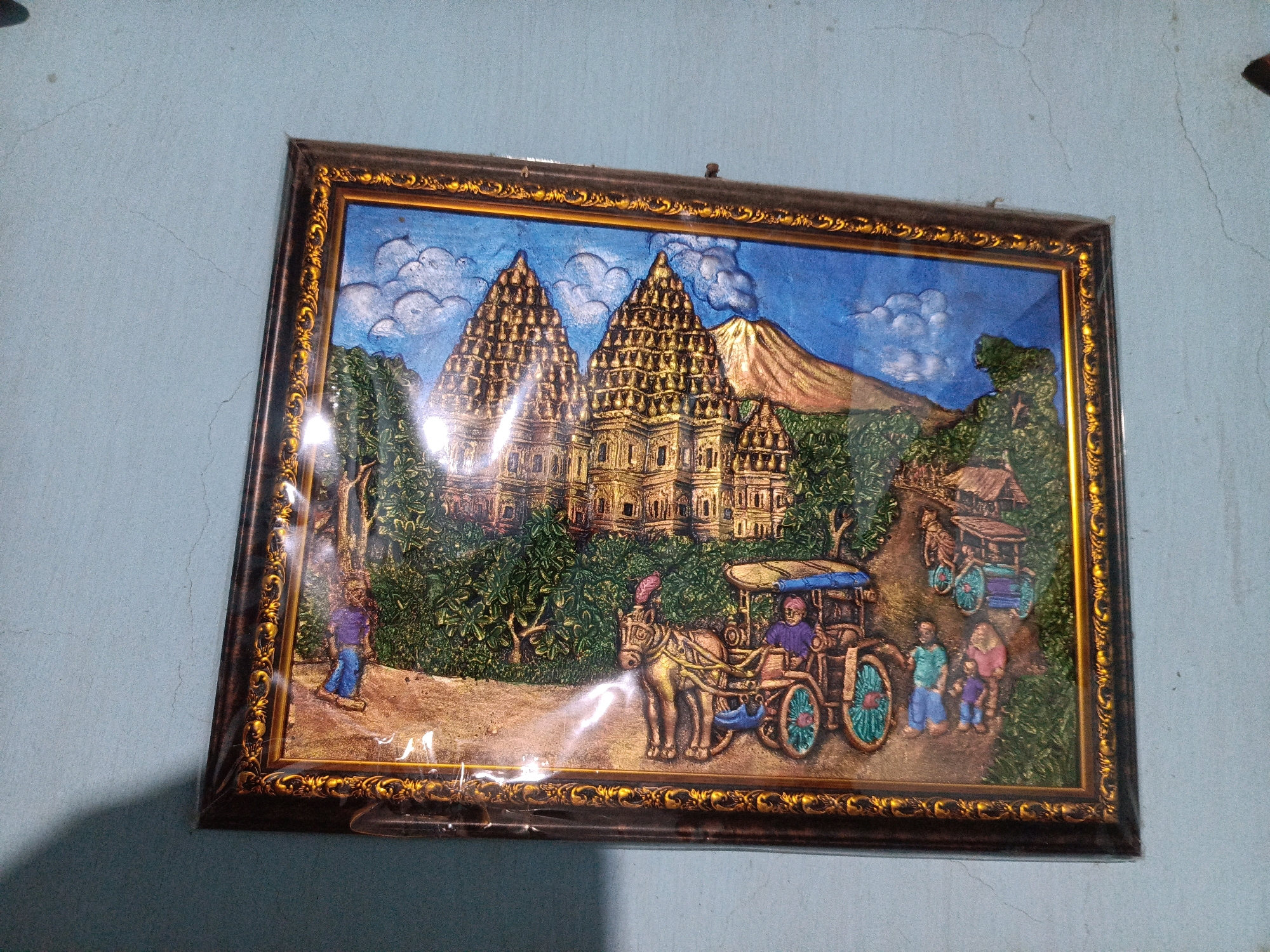 The image shows a framed, three-dimensional relief artwork hanging on a light blue, cracked wall. The artwork depicts a scene with several prominent elements. In the background, two large, ornate golden temples with tiered roofs are the central focus. To the right of the temples, a golden mountain peak rises against a bright blue sky with white clouds. The foreground of the artwork features lush green foliage and trees.
In the mid-ground, a horse-drawn carriage with a blue canopy is depicted, with a person driving it. The horse is a golden color and appears to be pulling the carriage. To the right of the carriage, there are several figures of people walking. Further in the distance, another smaller carriage or cart is visible. On the left side of the artwork, a lone figure in blue pants and a purple shirt stands near some trees. The overall scene appears to be a representation of a historical or cultural landscape, possibly inspired by Indonesian architecture and village life, given the context of Kalang, Indonesia. The artwork is encased in a dark brown and gold ornate frame, and the entire piece is covered in clear plastic, suggesting it is a decorative item.