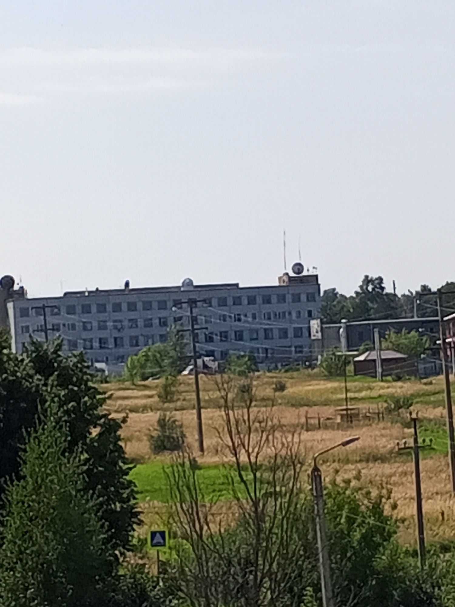 The image depicts an overcast day in Chistopol, Russia, with a long, multi-story building dominating the background. The building appears to be made of concrete and has numerous windows. Antennae are visible on the roof of the building, suggesting it may serve a communication or industrial purpose. In the foreground, there are trees, dry grass, and a street light. Power lines and poles are also present, indicating the presence of infrastructure. The overall scene suggests a somewhat industrial or utilitarian area on the outskirts of the city.