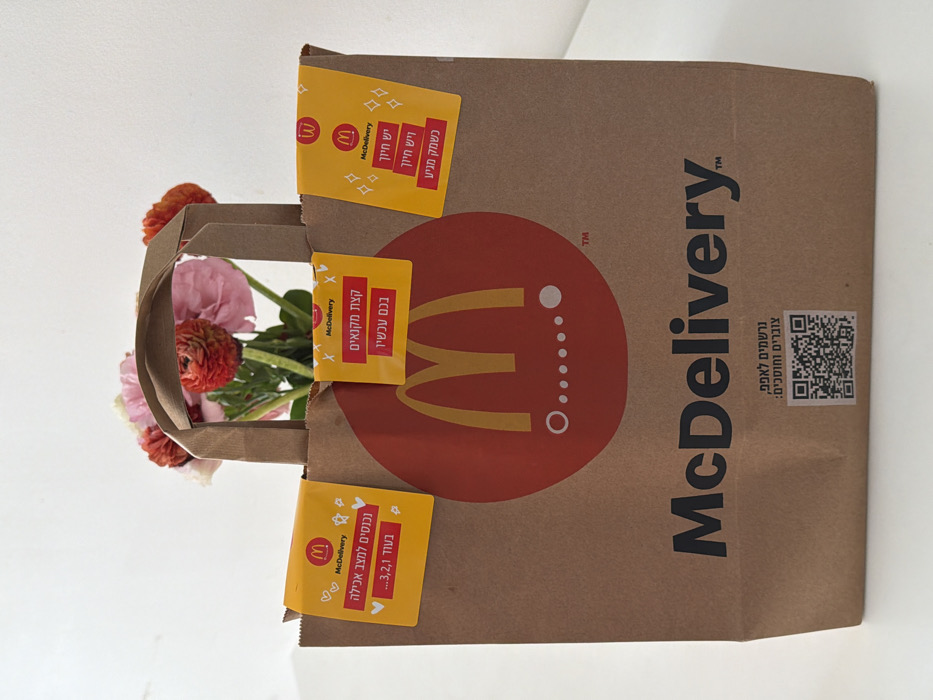 A brown paper McDelivery bag stands upright on a white surface. The bag prominently displays a large red circle with a yellow McDonald's "M" logo and a sequence of five white dots below it, four unfilled and one filled. Below this, "McDelivery" is printed in black text. Three yellow rectangular stickers are attached to the top edge of the bag. The left sticker displays a small McDonald's logo, "McDelivery" text, and the Hebrew phrase "נכנסים למצב אכילה 3,2,1..." (Entering eating mode 3,2,1...). The middle sticker has a small McDonald's logo, "McDelivery" text, and the Hebrew phrase "קצת מקנאים בכם עכשיו" (A bit jealous of you now). The right sticker also features a small McDonald's logo, "McDelivery" text, and the Hebrew phrase "יש חיוך כשתמק מגיע" (There's a smile when the Mc arrives). At the bottom of the bag, the Hebrew text "נרשמים לאפליקציה, צורפים וחוטפים:" (Register for the app, join and snatch/grab:) is printed above a black and white QR code. Partially visible within the bag is a bouquet of fresh flowers, featuring pink, red, and peach-colored blooms, possibly ranunculus. The background is an unadorned white wall or surface. The use of Hebrew text on the packaging indicates the location is in Israel, consistent with a scene in the city of Tel-Aviv.