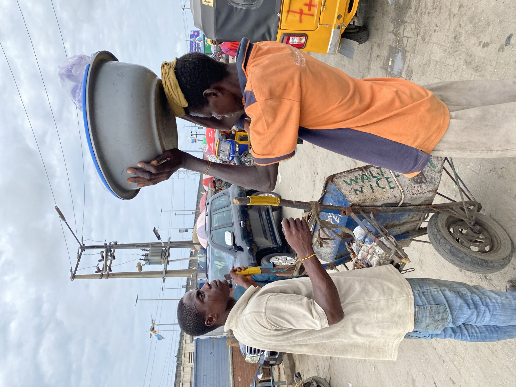 In the city of Lekki, Nigeria, the image shows two men in an outdoor setting, seemingly engaged in some form of roadside commerce or labor. The man on top is wearing an orange shirt with navy blue accents and light-colored pants. He has a yellow headband and is leaning his head on a large, gray, plastic bowl which is secured on top of a pushcart. The cart bears the words "WALT NICE." The man at the bottom is wearing a light tan shirt and blue jeans and is looking up at the first man while holding a yellow tool. Behind the men are several vehicles, including a yellow tricycle taxi, a dark SUV, and a variety of other cars. In the background, there are utility poles and buildings, indicating an urban setting. The sky is overcast.