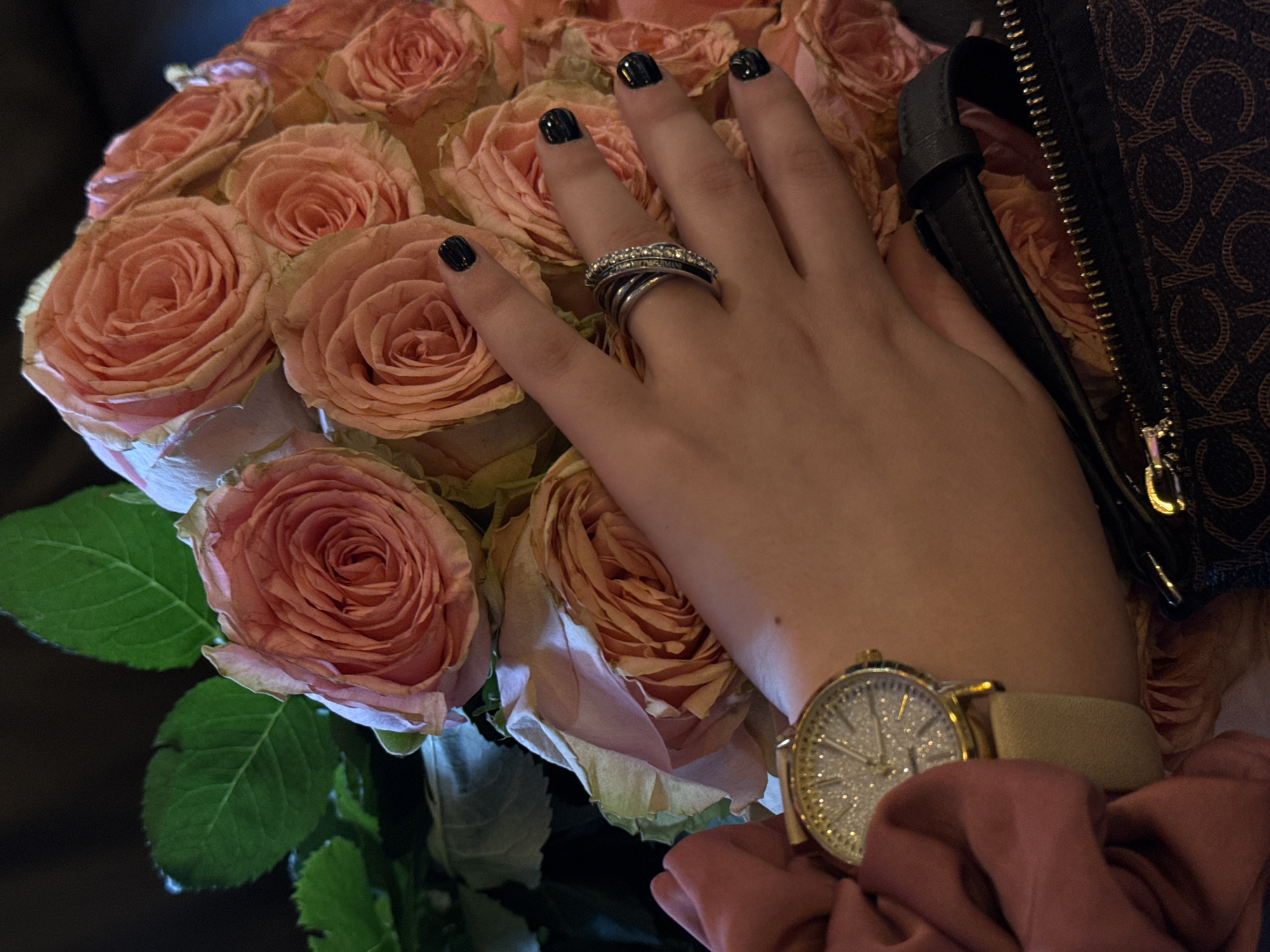 A person's hand rests on a bouquet of light pink roses. The hand has dark nail polish and wears a multi-band silver ring with stones. A gold-toned wristwatch with a light-colored band is visible on the wrist. The bouquet also supports a brown patterned bag with a zipper. A pink scrunchie is below the watch. There is no information in the image that indicates the location of this photo is in Yerevan, Armenia.