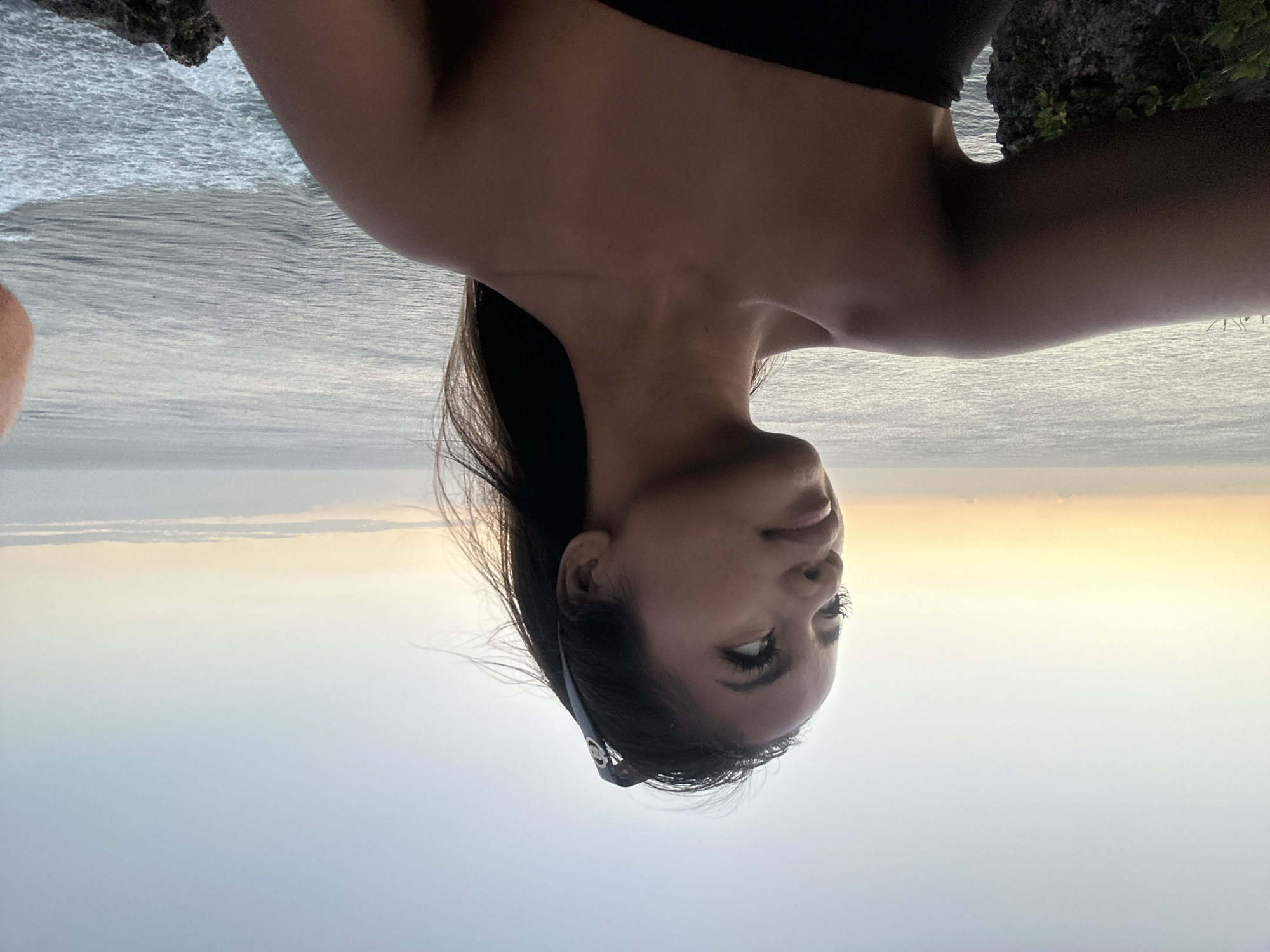 A young woman with long, dark hair pulled back by sunglasses poses for a selfie overlooking the ocean at sunset. She is wearing a black strapless top and stands on a cliff edge with lush vegetation visible below. The sky transitions from a pale orange at the horizon to a soft blue overhead, with a few wispy clouds. The ocean is calm with gentle waves breaking on the shore. The location is Kuta Selatan, Indonesia.