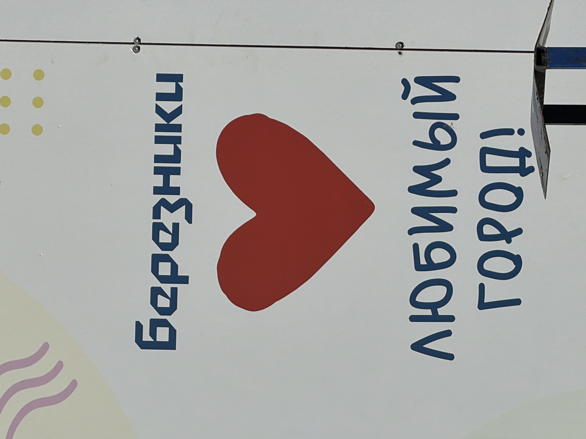 The image shows a sign in Berezniki, Russia, that reads "Березники ❤️ любимый город!", which translates to "Berezniki ❤️ favorite city!". The text is in blue. The heart is red. The background is white, with some pale yellow and purple abstract shapes on the left side. The sign appears to be made of a flat, rigid material. A piece of a metal support structure is visible on the upper right side of the image. The sign is mounted with rivets in the upper portion of the frame.