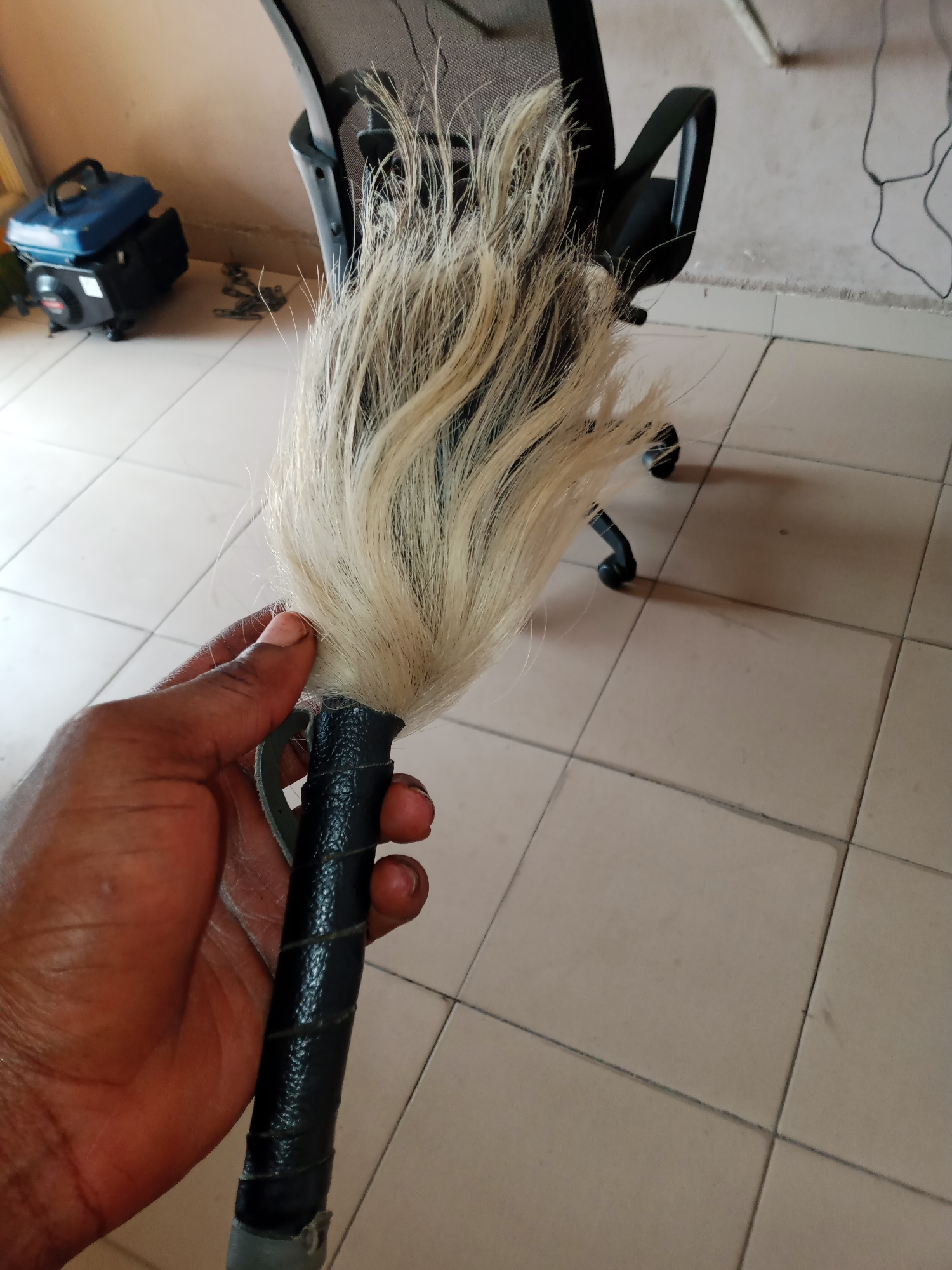 A hand with dark skin holds a whisk. The whisk has a black handle wrapped with a textured material, and the whisk itself is made of blonde hair. Behind the hand and whisk, a portion of a black office chair with a mesh back is visible. To the left of the chair, a small, blue and black portable generator is on the tiled floor. The floor is made of large, square, beige tiles. The background wall is a light, neutral color, and electrical cords are visible hanging down it. The scene appears to be indoors, possibly in a home or office. The lighting suggests it is daytime. There is no visible text in the image.