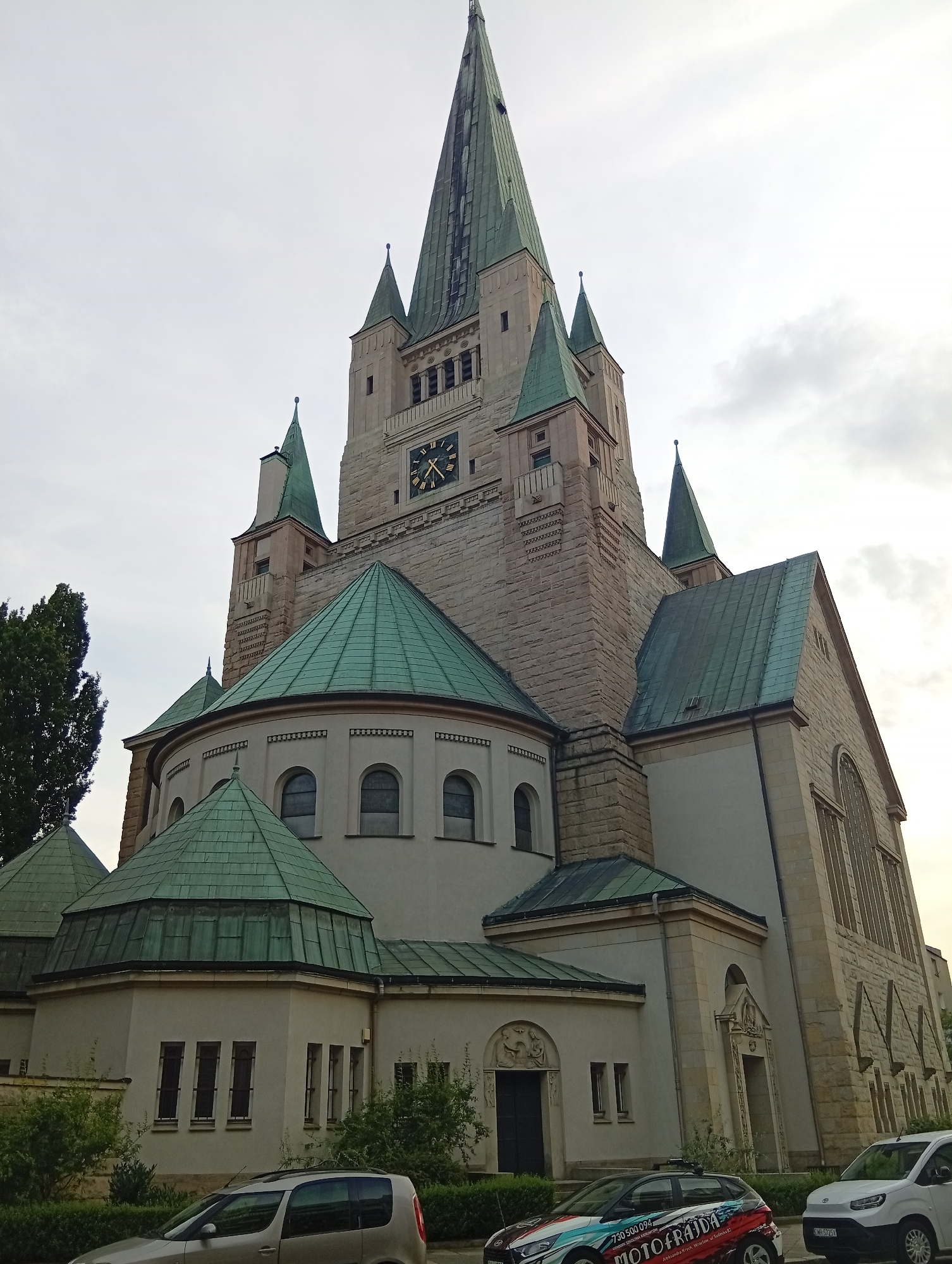 The image captures an external view of the Church of the Divine Mercy in Wrocław, Poland. The church features a prominent clock tower with Roman numerals and a green spire, surrounded by smaller green-roofed turrets. The building's facade is constructed of light-colored stone blocks. In the foreground, two cars are parked on the street; the one on the left is a silver hatchback, and the one on the right is a white van. The sky above is overcast.
