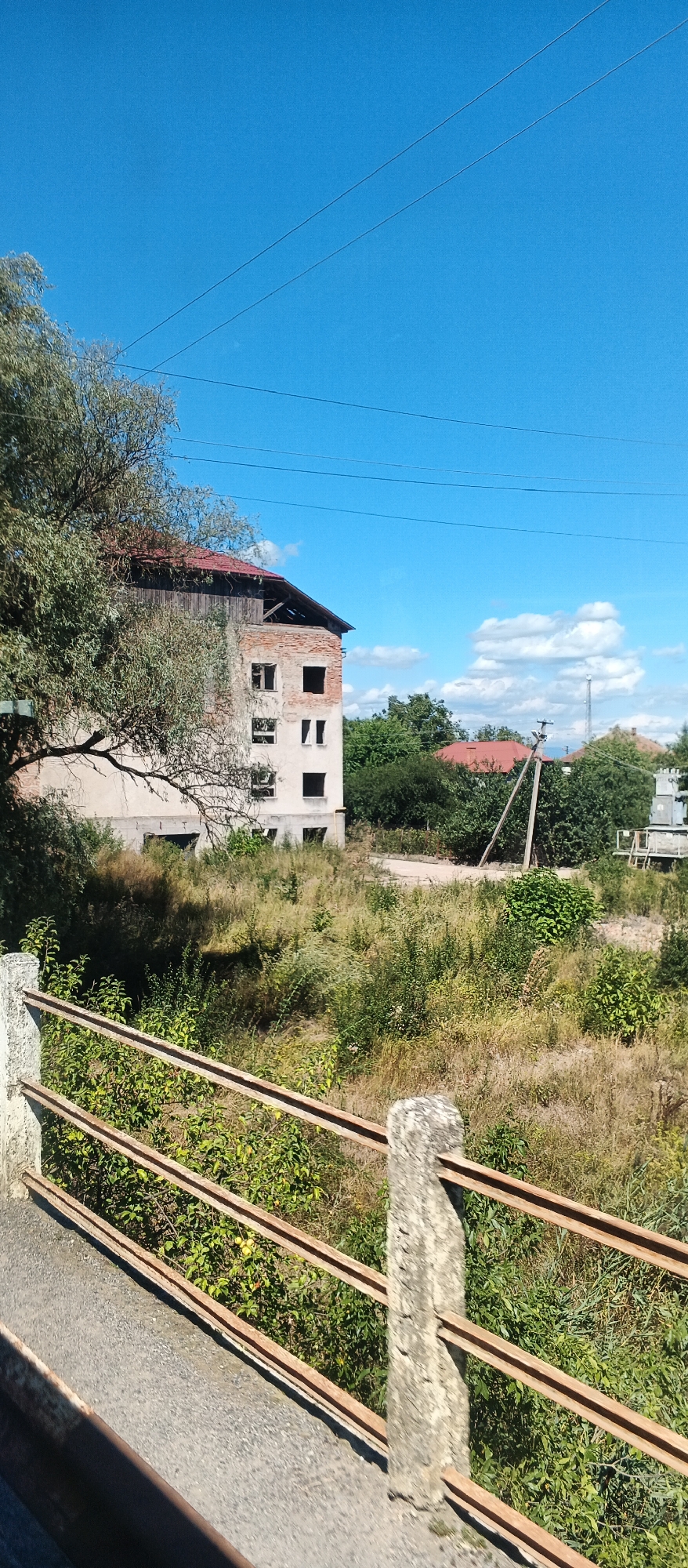 The image shows an unfinished building with exposed brickwork and empty window frames. The building has a red tiled roof with some sections missing, revealing wooden beams underneath. The exterior walls are a light color, and some areas appear to be plastered, while others are bare brick. In the foreground, there is a rusty metal railing supported by concrete posts. This railing runs along what appears to be a pathway or bridge. The area surrounding the building is overgrown with tall, dry grass and various green shrubs and trees. In the background, more trees and other buildings with red roofs are visible. The sky is a bright, clear blue with a few scattered white clouds. There are power lines running across the upper portion of the image. The scene appears to be an outdoor environment in daylight.