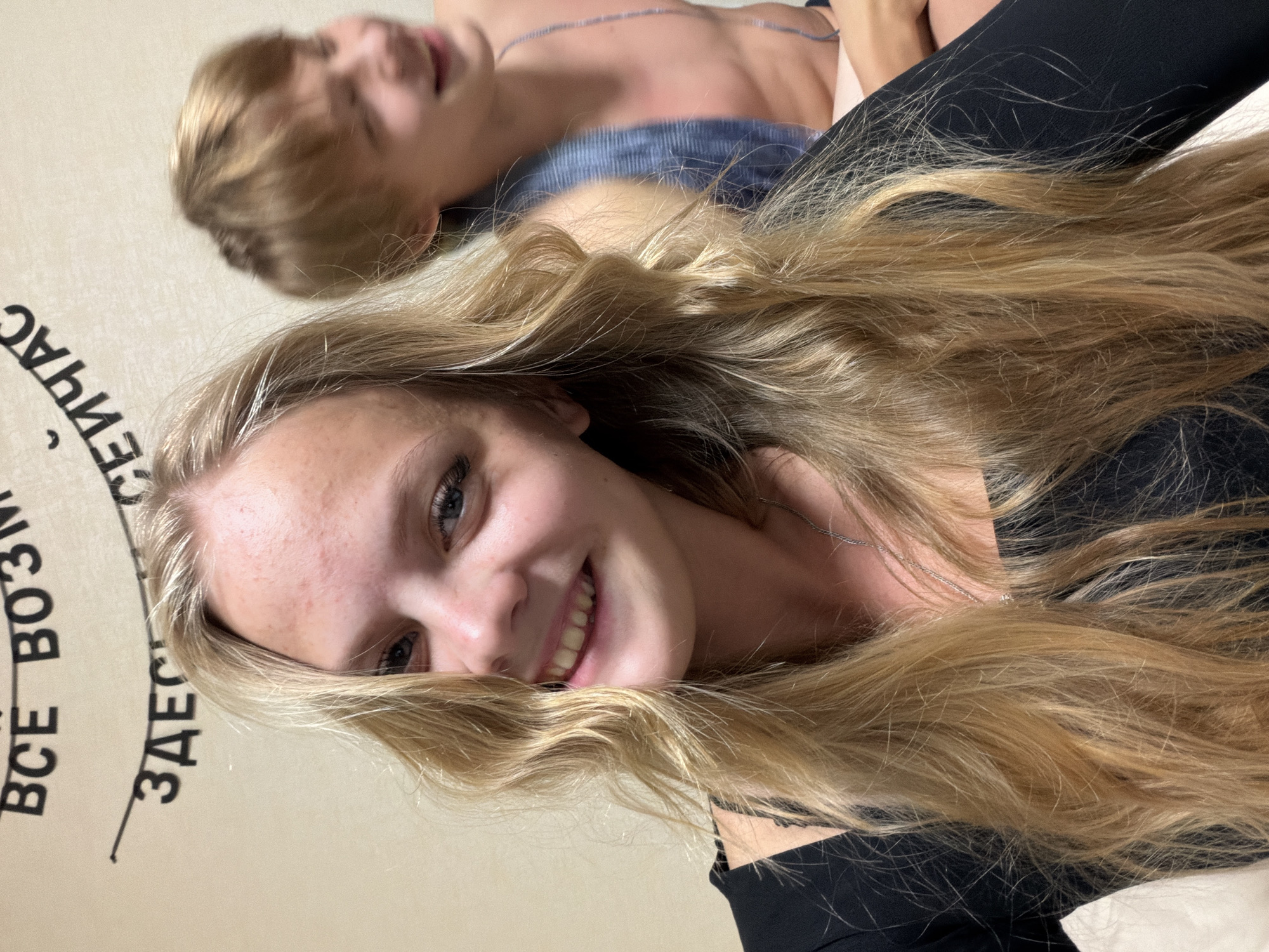 The image shows a young Caucasian female with long blonde hair, wearing a black top, smiling at the camera. Behind her, a young Caucasian male with blonde hair is visible, also smiling and looking to the side. The background features a wall with black lettering in Russian, part of which reads "ВСЕ ВОЗМОЖНО" (Everything is possible) and "ЗДЕСЬ И СЕЙЧАС" (Here and now). The setting appears to be an indoor environment. The presence of Russian text suggests the location could be Russia, and specifically the city of Izhevsk, though this cannot be definitively confirmed from the image alone.