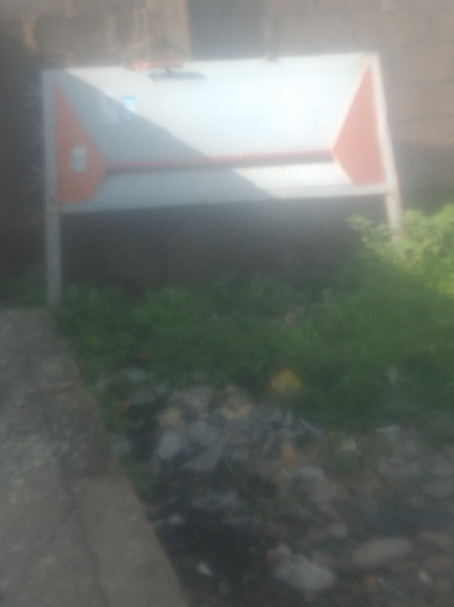 The image depicts an outdoor scene in Jalingo, Nigeria. In the foreground, a narrow, paved pathway runs diagonally from the bottom left to the center of the frame. To the right of the pathway, a patch of overgrown green vegetation is visible, interspersed with scattered debris, including plastic and other refuse.
Dominating the mid-ground is a large, rectangular object that appears to be a sign or a display board. It is predominantly white with a prominent orange triangle on the left side and another orange triangle on the right. The board is supported by thin, light-colored vertical poles.
The background is blurred, showing a rough, textured wall or building surface. The overall impression is that of a cluttered urban or semi-urban environment. The lighting suggests it is daytime, but the image quality is poor, with significant blurriness. No people are discernible in the image.