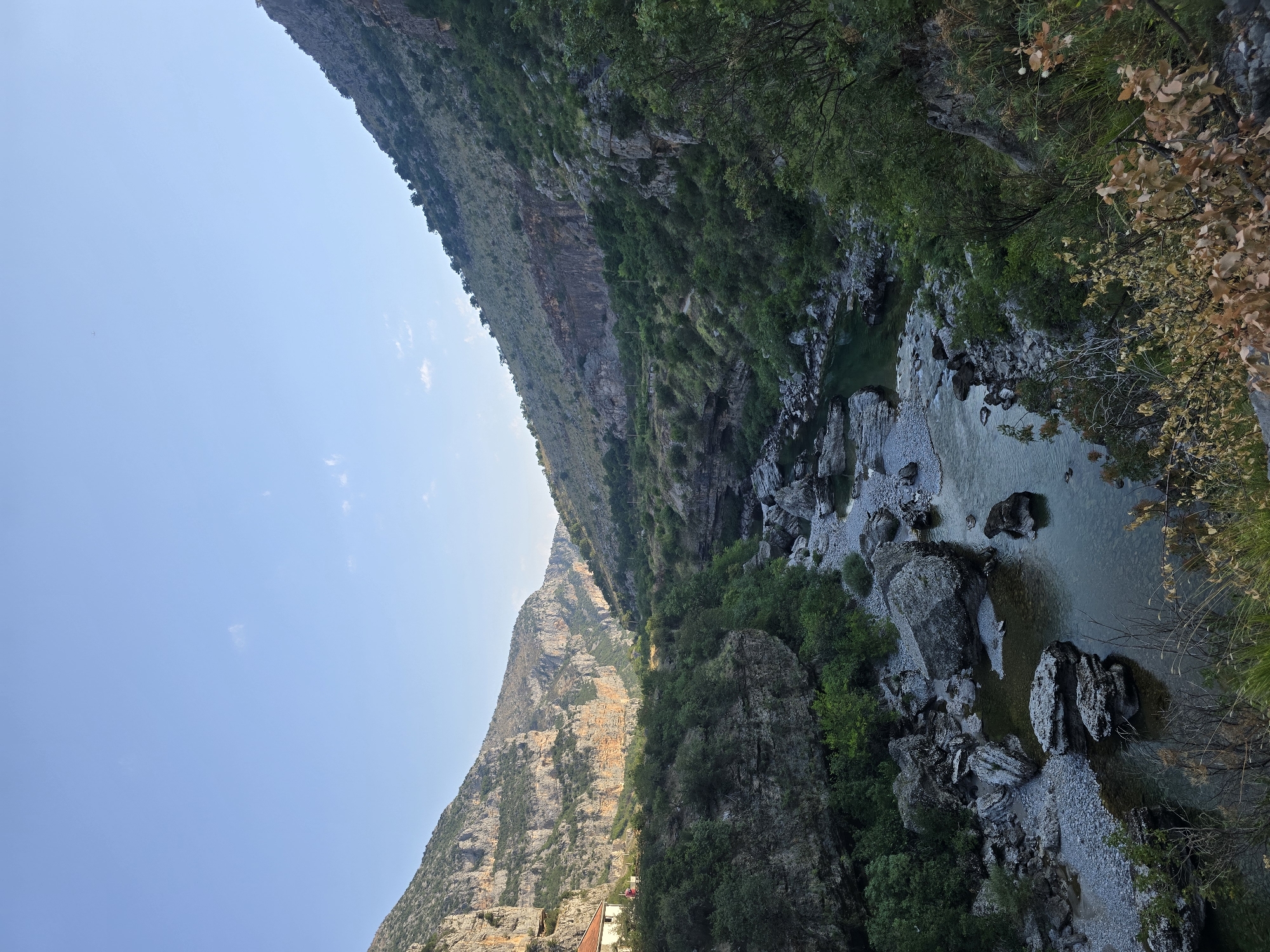 The image depicts a canyon with a river flowing through it. Steep, rocky mountains flank the canyon, covered in patches of green vegetation. The river is clear and shallow in some areas, with large rocks visible along its bed. On the left side of the canyon, a white building with a red roof is situated on a ledge overlooking the river. The sky is a clear, pale blue. The scene appears to be in a natural, undeveloped area.