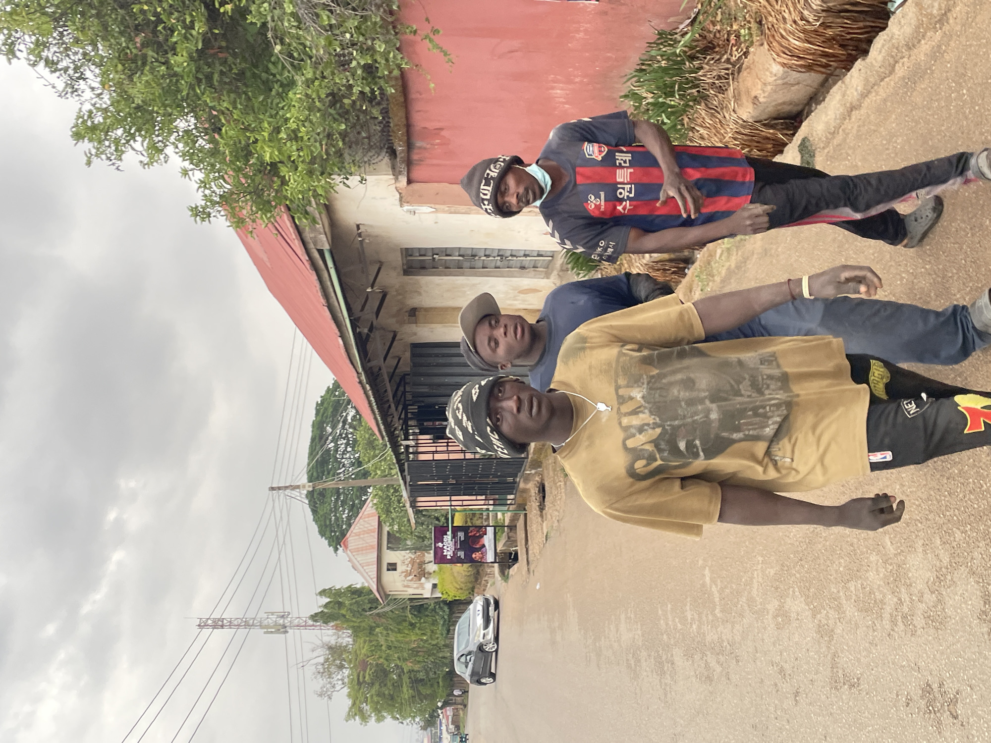 Three young men are positioned closely together on a paved road or sidewalk. The first man, closest to the camera, wears a khaki or beige colored shirt and black pants with yellow and red flames on the left leg, along with a patterned beanie. The second man is clad in a navy blue shirt and gray pants, and wears a gray baseball cap. The third man wears a soccer jersey with blue and red stripes and a black beanie, also with a surgical mask hanging loosely from his ear. Behind them is a building with a red roof and a dull red wall, and vegetation hangs over part of the building. Across the road, there is a billboard and several vehicles driving on the road. Power lines are visible against a cloudy sky. The scene is located in Jos, Nigeria.
