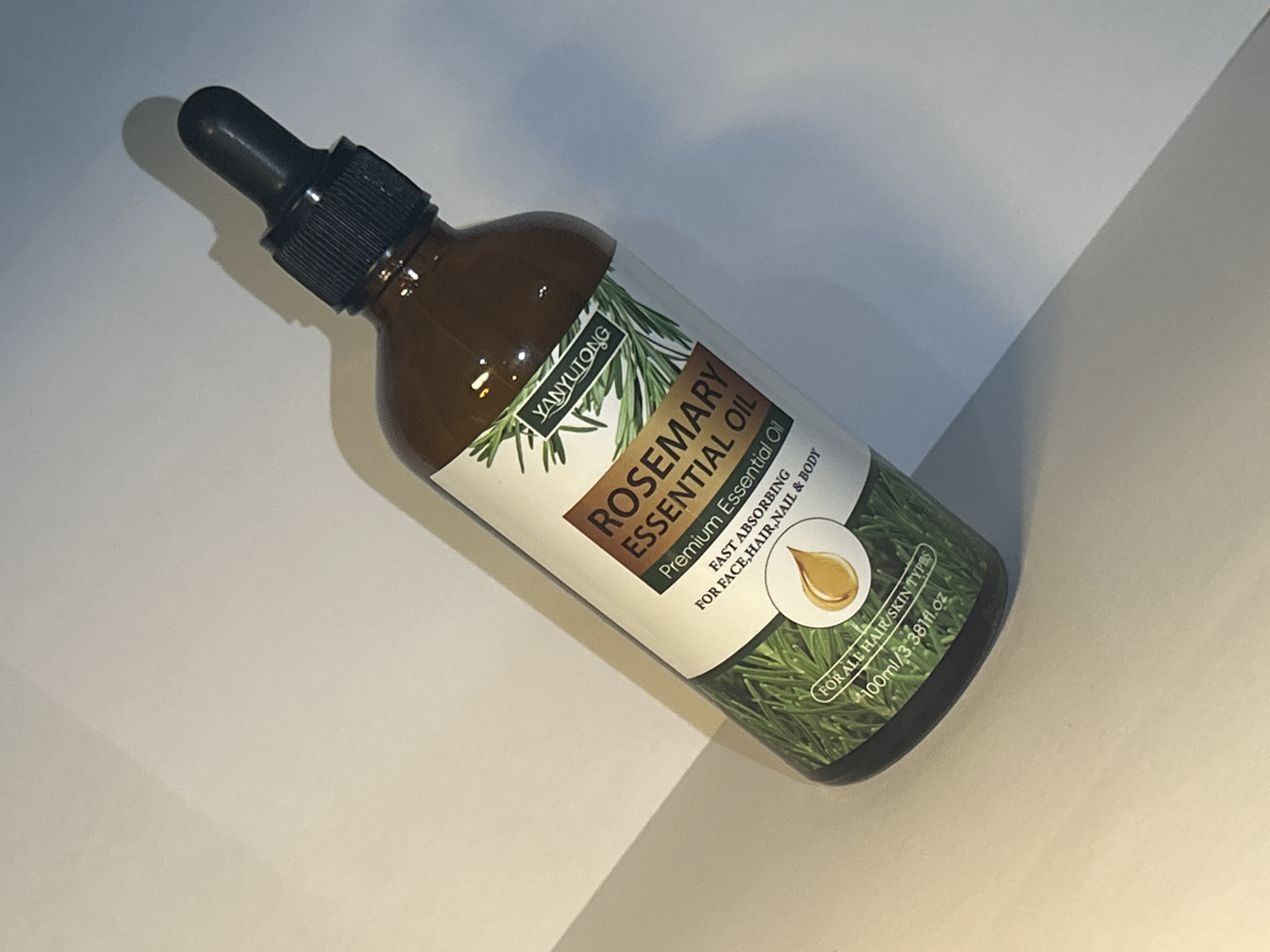 The image shows a bottle of Yanyutong Rosemary Essential Oil. The bottle is made of dark brown glass and has a black dropper lid. The label is white with green accents, featuring an illustration of rosemary sprigs. Text on the label indicates the product is "Premium Essential Oil," "FAST ABSORBING," and suitable "FOR FACE, HAIR, NAIL & BODY." It also states "FOR ALL HAIR/SKIN TYPES" and provides the volume as "100ml/3.38fl.oz." The bottle is placed on a white surface, possibly a table or desk, with a diagonal shadow cast across it, suggesting the lighting source is from the side. The background is a plain, light-colored surface, with a distinct line where the surface meets another, creating a shadowed area. There are no people visible in the image. The scene appears to be an indoor product shot, likely for commercial purposes, with the focus solely on the oil bottle. The lighting is bright and casts a distinct shadow, indicating it is likely artificial light. No specific location cues within Yizhivtsi, Ukraine, are discernible from the image itself.