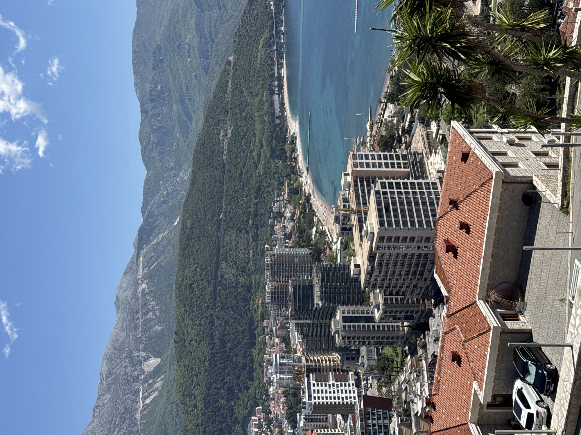 The image shows a coastal city with a backdrop of mountains. The city features numerous buildings, including high-rise structures with modern architectural designs, interspersed with older structures featuring terracotta roofs. A sandy beach runs along the coastline, bordering the clear blue sea. A dense forest stretches up the mountainside, contrasting with the bare rock faces of the higher elevations. Two cars, one white and one black, are parked on a paved area next to a building with a red-tiled roof. The sky is clear with a few scattered clouds.