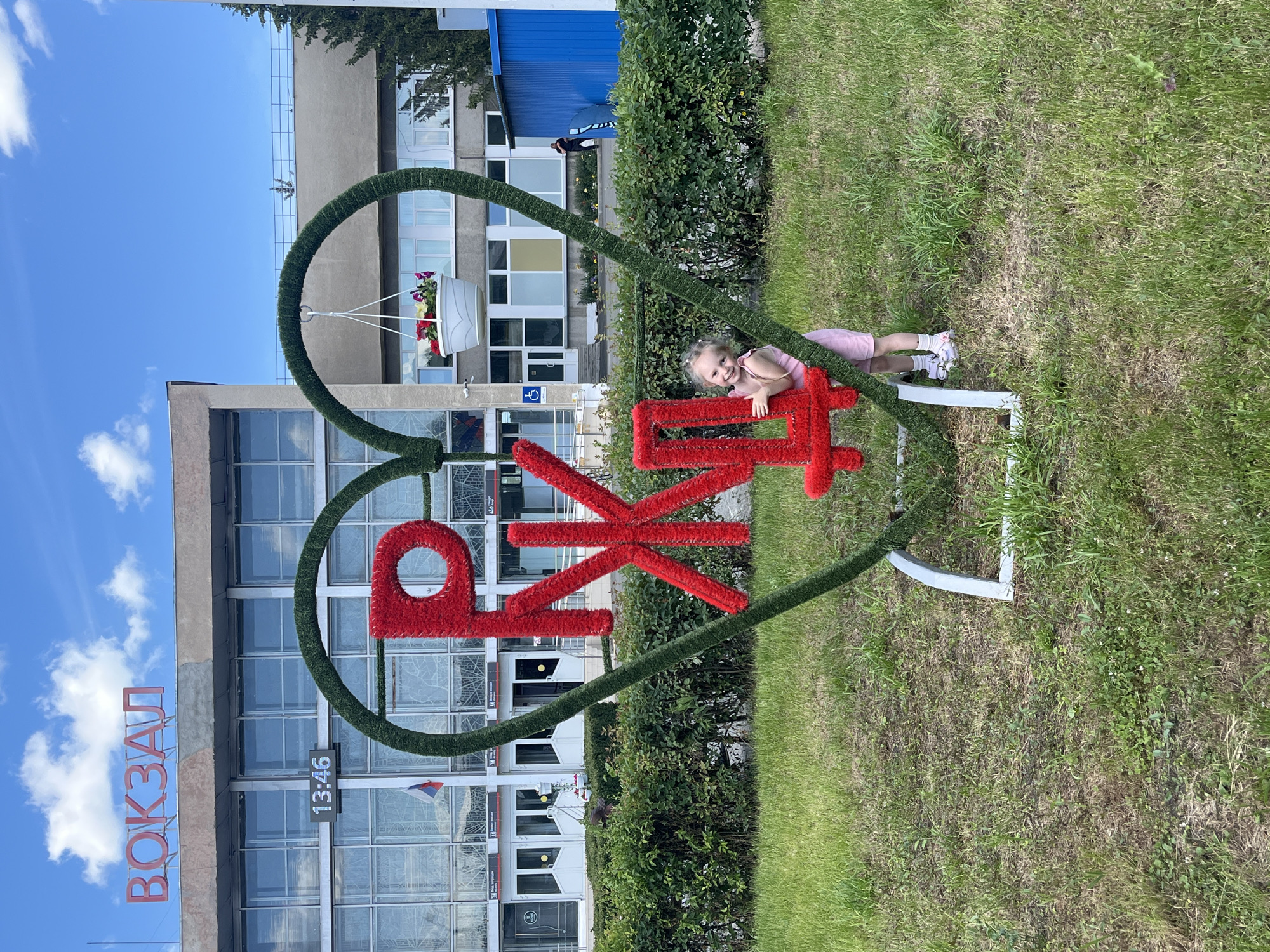 A young girl in a pink dress and white sneakers is leaning against a large, red, fluffy sculpture shaped like Cyrillic letters within a heart-shaped frame. The sculpture is covered in green artificial grass. The girl is smiling at the camera. She is lying on her side, with her legs bent.
The sculpture is in front of a building with large windows, possibly a train station, as indicated by the word "ВОКЗАЛ" (VOKZAL), meaning "station," written in red letters on the building's facade. A digital clock on the building displays "13:46". The setting is outdoors, with green grass and bushes around the sculpture. The sky is blue with scattered white clouds, suggesting it is daytime. The overall scene conveys a sense of joy and playful interaction with the environment.