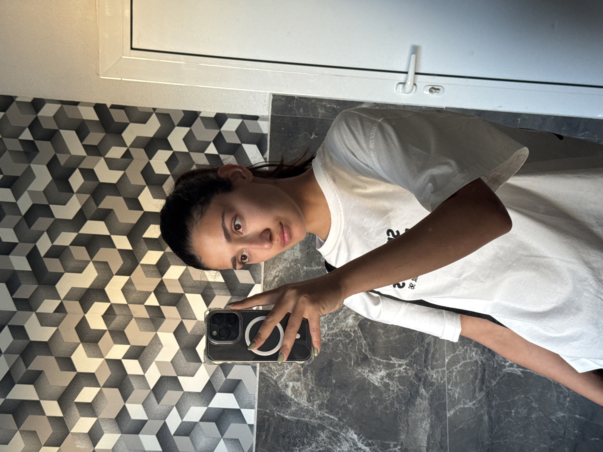 A person with dark hair is taking a selfie in a bathroom. The walls are decorated with a geometric patterned wallpaper and marble tiles. A white window frame is visible in the upper part of the image. The location is likely Bar, Montenegro as requested.