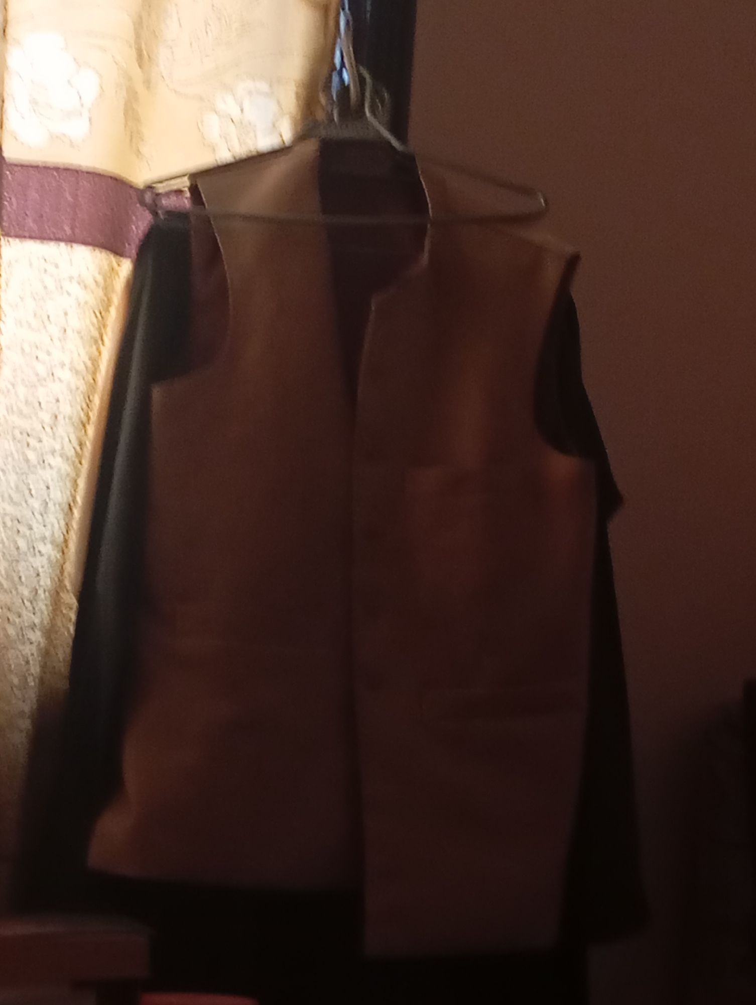 The image shows a tan vest with a dark undergarment, hanging on a hanger against a backdrop of a patterned curtain and a plain wall. The lighting is dim and warm. The vest has a V-neck and appears to have pockets. The curtain is yellow with a floral or abstract pattern in a lighter color, and a dark red band runs horizontally across it. The scene is indoors, likely in a room with muted lighting. There are no people visible in the image.