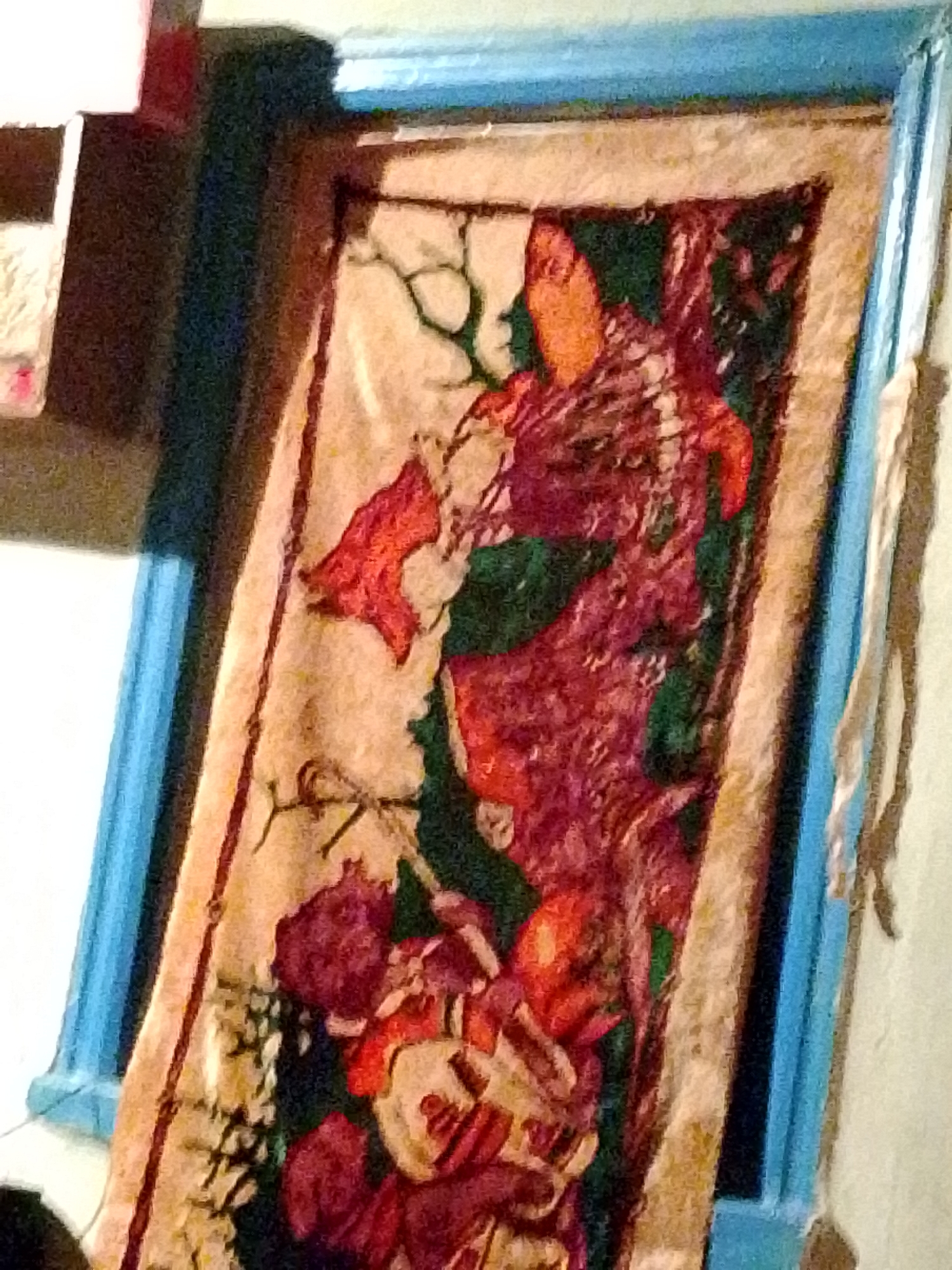 A vertically oriented fabric artwork, possibly a tapestry or batik, is depicted within a light blue structural frame, rotated approximately 15-20 degrees clockwise. The artwork features abstract or stylized organic forms in shades of red (maroon, orange-red) and dark green against a light beige background. The upper section displays an orange-red shape with intricate reddish patterns extending downwards, alongside dark, thin, branch-like elements. The middle and lower sections are dominated by dense reddish and maroon patterns interspersed with dark green, including shapes that resemble stylized fruits or clustered forms. The light blue frame borders the artwork on the top, right, and bottom. To the left of the artwork, a dark vertical element and a partial view of a light-colored, possibly white, horizontal surface with a red underside are visible. To the right of the blue frame, a light-colored wall is present, with a faint string or cord hanging from the upper right. The image quality is grainy, suggesting low-light conditions. Location: Unknown, Unknown.