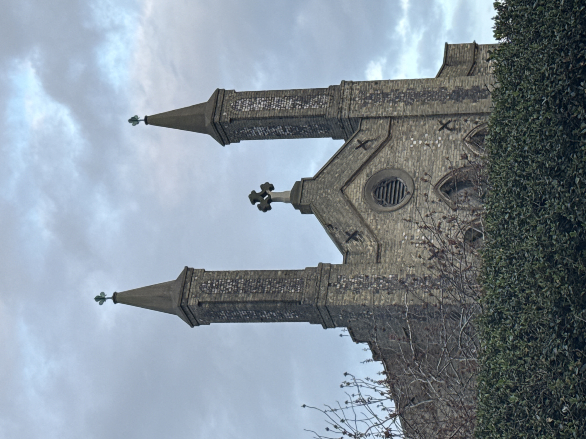 A low-angle, medium shot depicts a section of the St. Mary Magdalene Church in Great Yarmouth, United Kingdom. The architecture is Gothic Revival style, evidenced by the presence of two towers with pointed spires topped with green, clover-shaped ornaments. A third, shorter tower rises between them, crowned with a cross. The towers are constructed of light-colored brick or stone, with darker detailing. A circular window is visible in the main facade. A dense green bush obscures part of the church's lower right portion. Bare branches extend from the bush towards the brickwork. The sky overhead is overcast with grey and white clouds.
