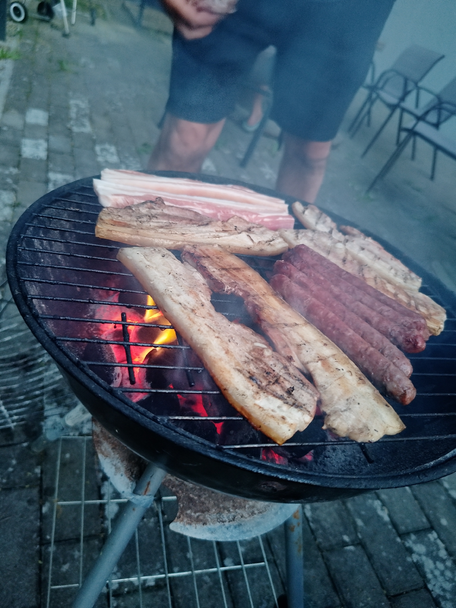 A medium shot depicts a black round charcoal grill, heavily laden with food being cooked over hot coals, situated on a brick-paved surface in an outdoor setting. The food includes several strips of bacon, pork belly, and sausages. Behind the grill, a man stands with bare legs wearing dark shorts. Several chairs are visible in the background. The location and city are unknown.