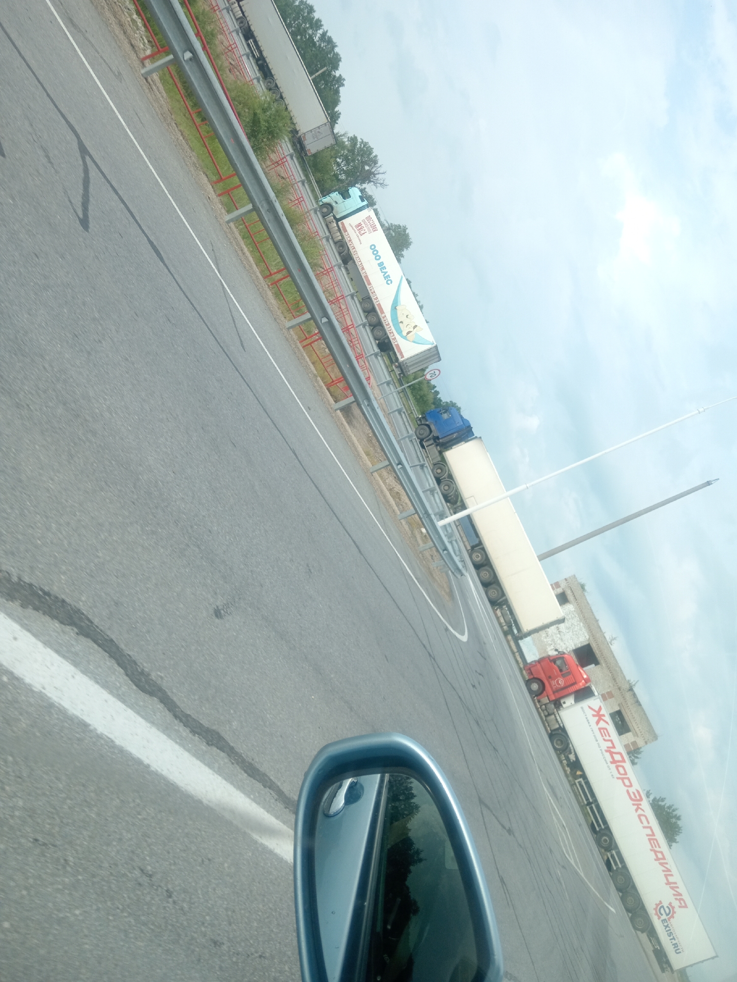The image shows a perspective from a vehicle, looking towards a highway with several trucks. The road is asphalt with white lines, and there is a metal guardrail on the side. A blue truck with a white trailer is visible, displaying text in Russian. Another white trailer truck with red lettering, also in Russian, is parked further down. A red truck cab is attached to this trailer. In the background, there is a building with a brick facade and some trees. The sky is cloudy. The angle of the shot suggests the vehicle is moving, and the mirror on the right side of the frame confirms this. The presence of the trucks and the highway suggest a commercial or industrial area. Based on the visible Cyrillic text indicating a Russian company, and the road infrastructure, this scene could be located near Vasil'evka, Russia, a common location for such commercial transport hubs.