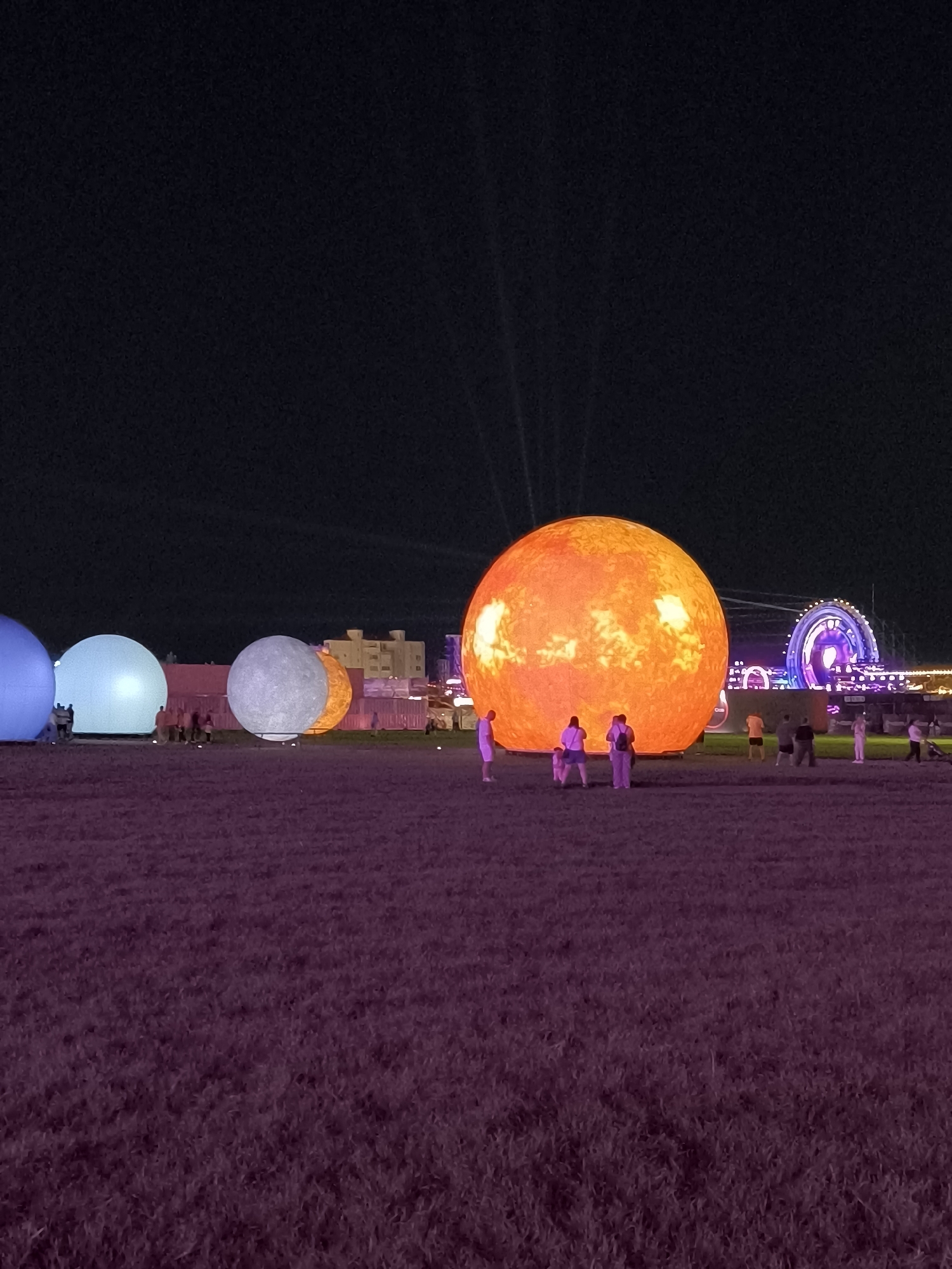 The image captures an outdoor nighttime scene at the "Living Planet" exhibition in Costinești, Romania, featuring large inflatable planetary models. In the foreground, a field of dry grass is illuminated with a soft purple light. Dominating the midground is a massive, bright orange inflatable sphere representing the sun, with intricate surface details. To its left, a textured, grey inflatable sphere, likely the moon, is partially visible. Further to the left, two more large, softly glowing spheres, one blue and one light blue, are seen. Several individuals are scattered around the inflatable models, observing them. In the background, buildings with illuminated windows are visible, and a large, brightly lit Ferris wheel with colorful purple and white lights is prominently displayed. The sky above is dark and clear.