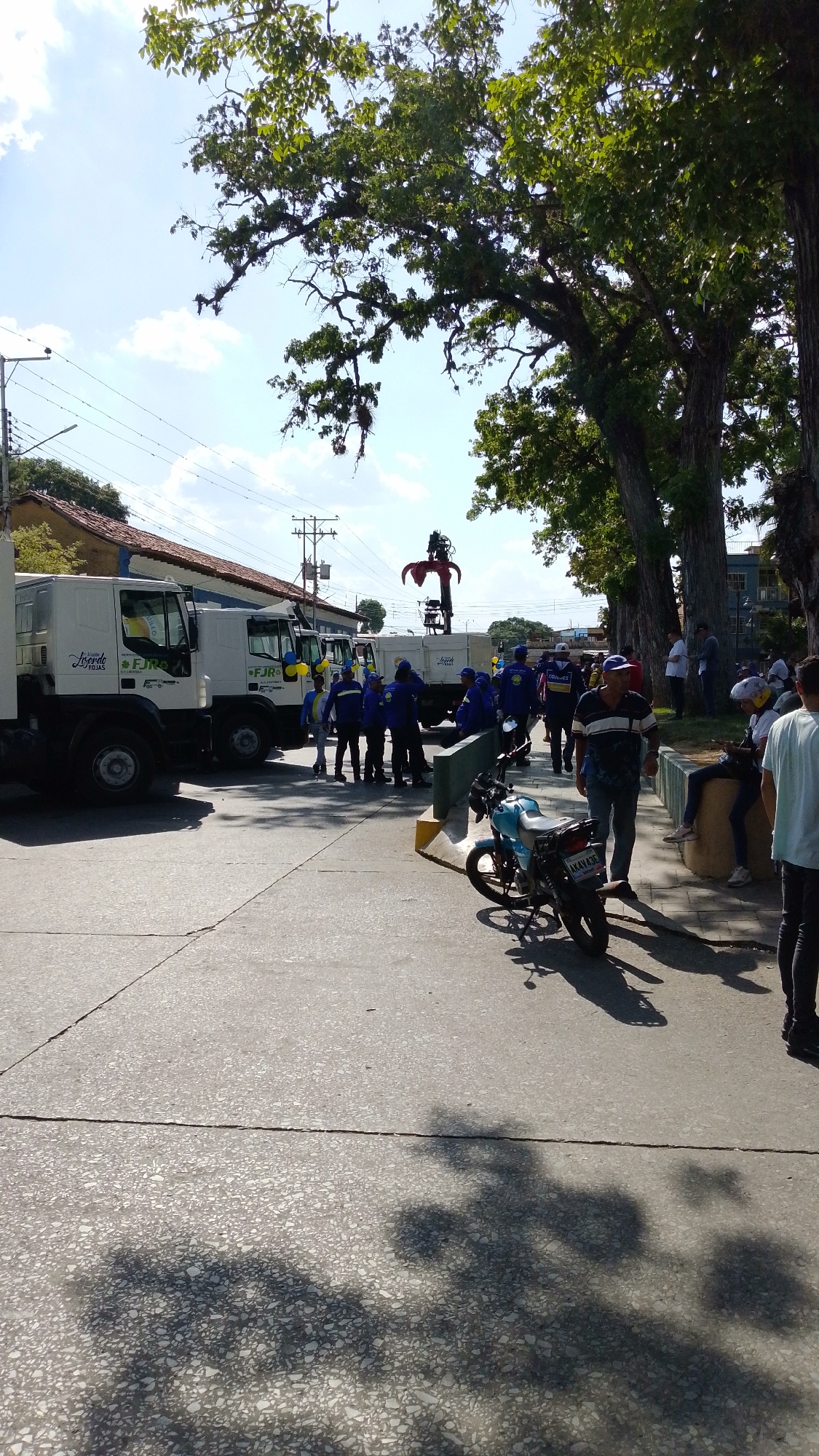 The image shows a sunny day in Tinaquillo, Venezuela. A row of white utility trucks with the "FJR" logo are parked along a street. A group of people, most wearing blue uniforms, are gathered near the trucks. A blue motorcycle is parked nearby. People are sitting on a low concrete wall and walking. Large trees with visible shadows are present.