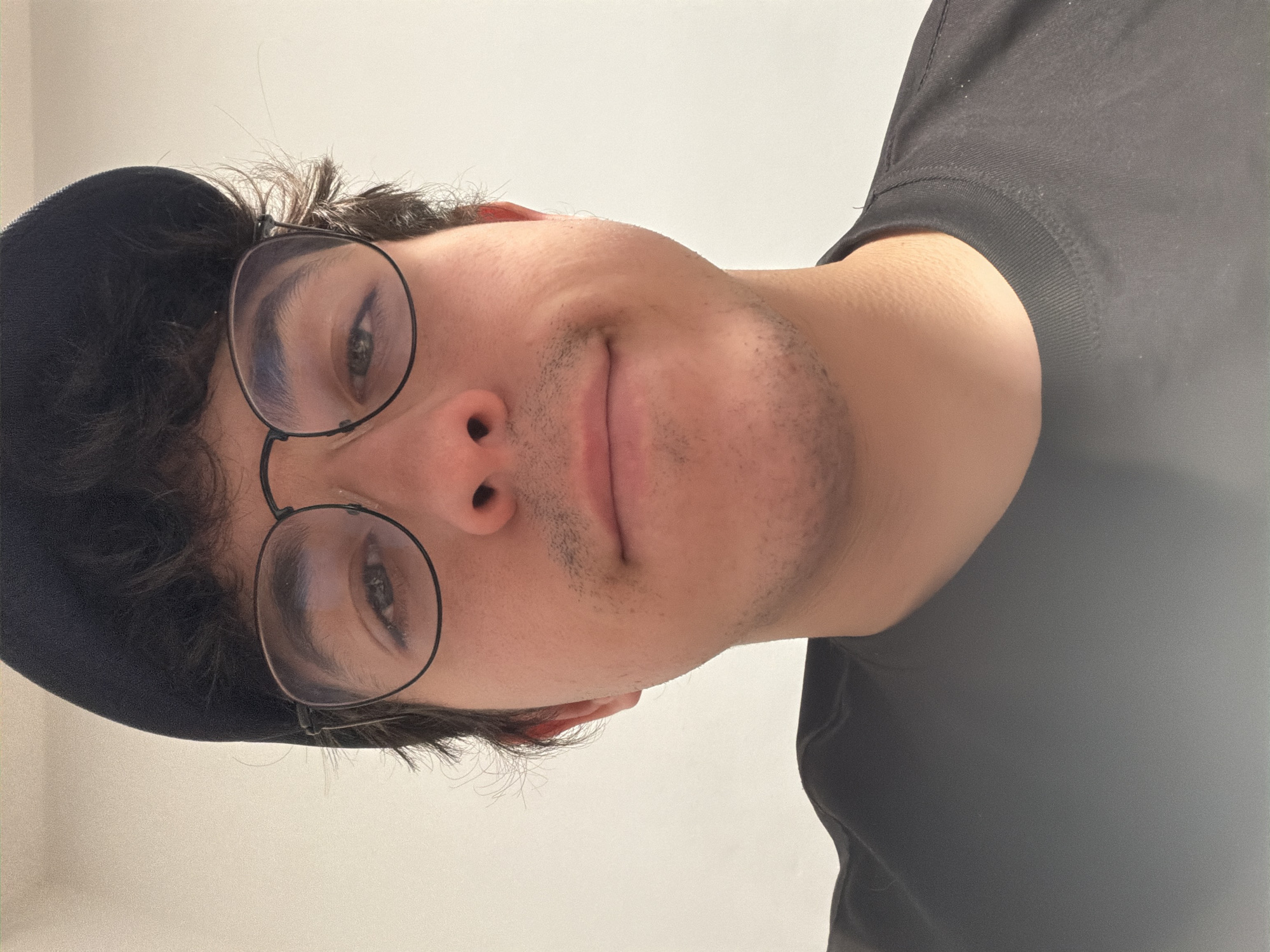 The media file is a close-up, vertically oriented, selfie-style photograph of a young person wearing a black t-shirt and a black baseball cap. The person is wearing round-framed glasses and has dark, curly hair. There are some light stubble markings on their chin and upper lip. The lighting suggests an indoor setting with a soft, diffused light source, possibly from a window or artificial lighting. The background is a plain, light-colored wall. The person's expression is neutral, with their eyes looking towards the camera. The overall impression is casual and informal. There is no visible text or discernible weather or time of day cues, beyond the general lighting. The location context of Cancún, Mexico, is provided separately and is not evident from the visual content of the image itself.