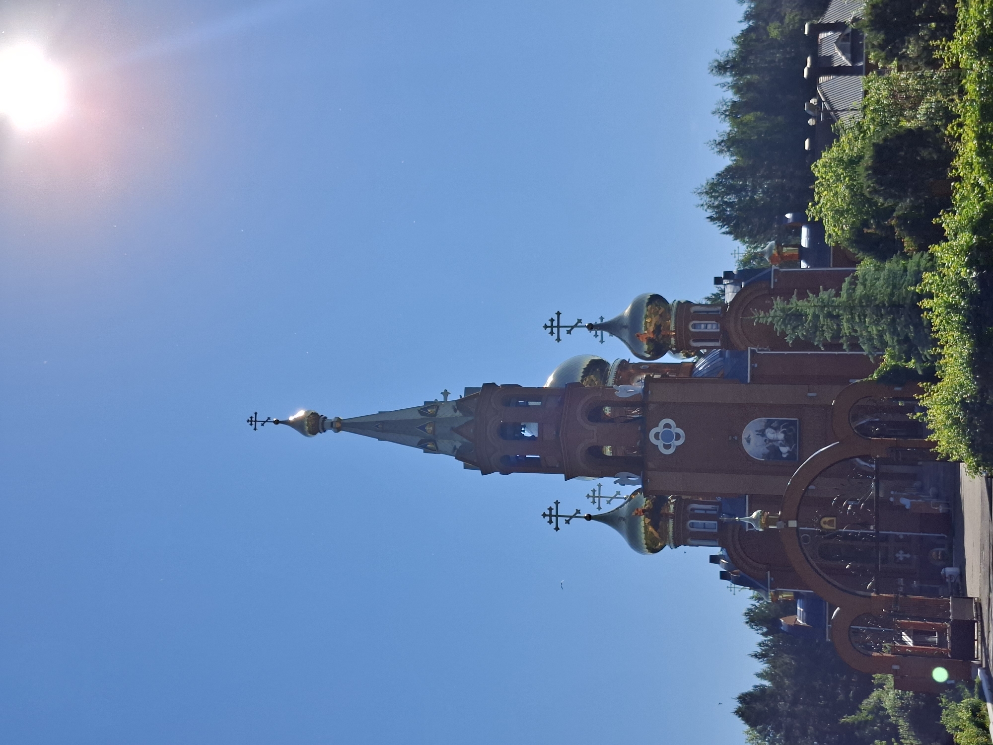 An exterior, low-angle, full shot captures a brick Orthodox church against a clear, bright blue sky. The sun is visible in the upper right corner, casting a bright glare. The church has multiple golden domes, each topped with a cross. A tall, slender tower rises from the center, also adorned with a cross. The building features arched entrances with ornate metal gates. Green trees and foliage surround the church, with some buildings visible in the background behind the trees. The scene appears to be in a natural setting, possibly a monastery or a church on the outskirts of a city.